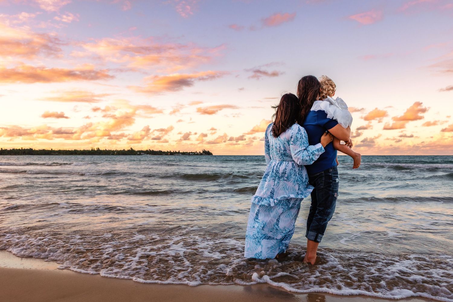 Family of three watching a stunning sunset on a tropical beach, waves gently washing over their feet.