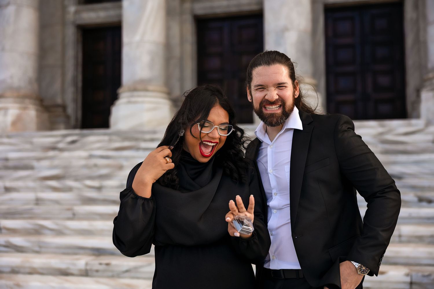 Couple in black attire smiling and posing on steps of a grand building.