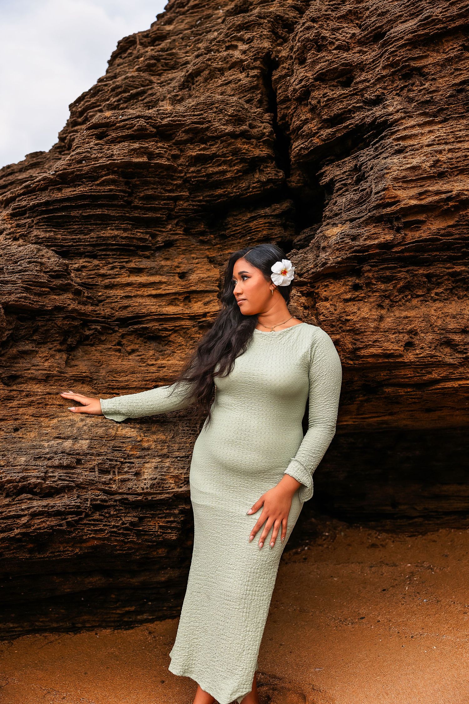 Pregnant woman in sage green knit dress with white flower in hair poses against red rock formation outdoors.