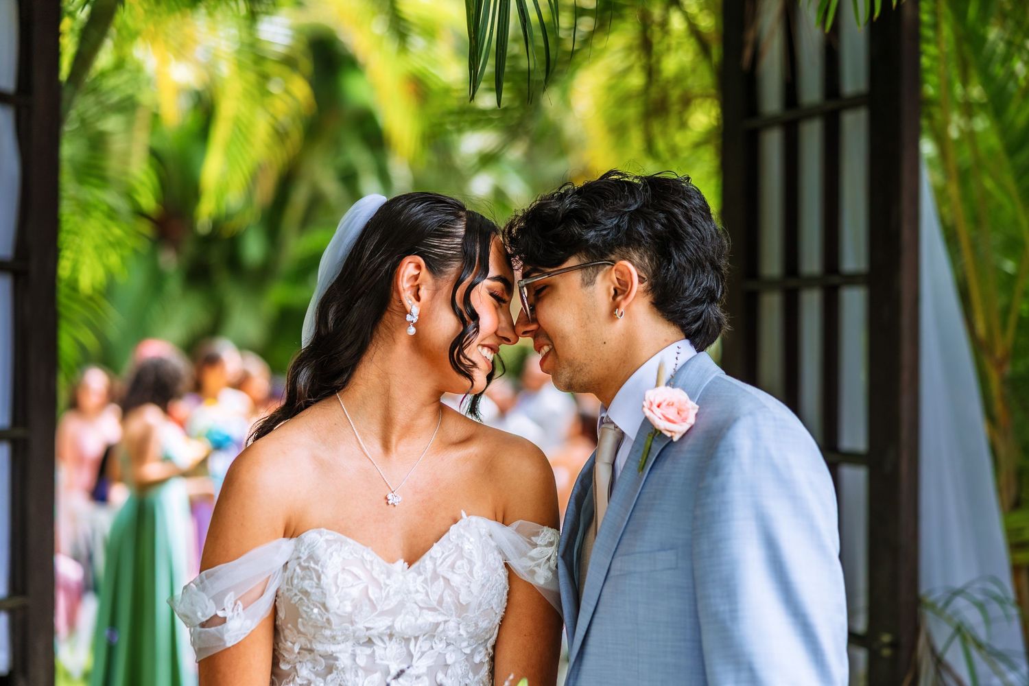 Bride and groom sharing an intimate moment, touching foreheads at an outdoor wedding ceremony surrounded by tropical greenery.