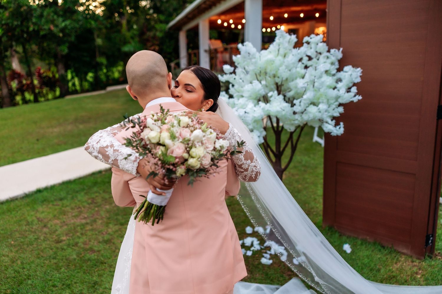 Bride in white gown with veil embraces guest in pink dress outdoors at wedding venue with floral bouquet.