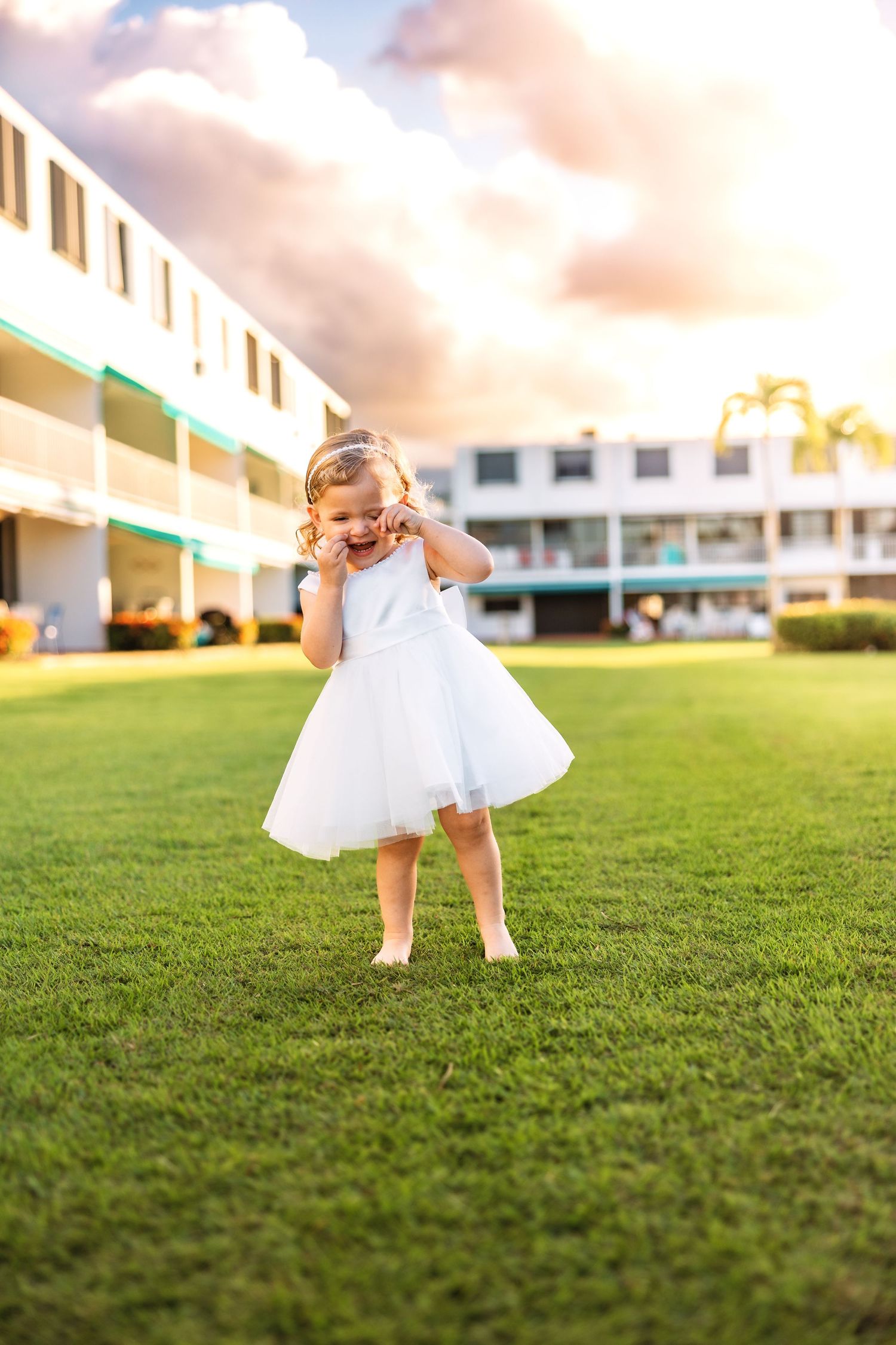Joyful toddler girl in white tulle dress laughing on green lawn at golden hour near a resort building.