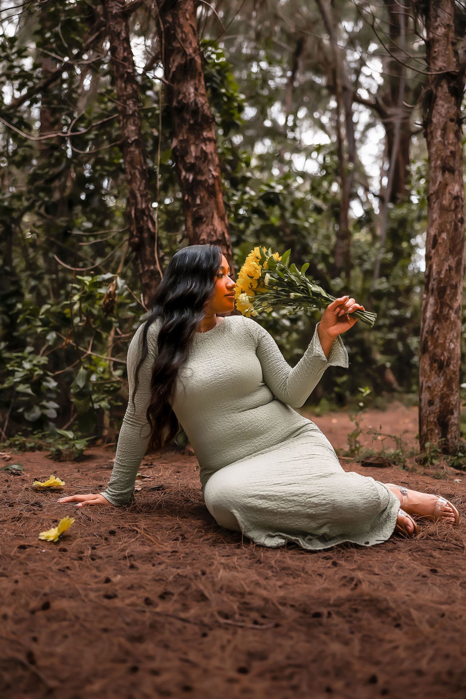 A woman in a sage green dress sits on forest ground, holding yellow flowers, surrounded by tall trees.