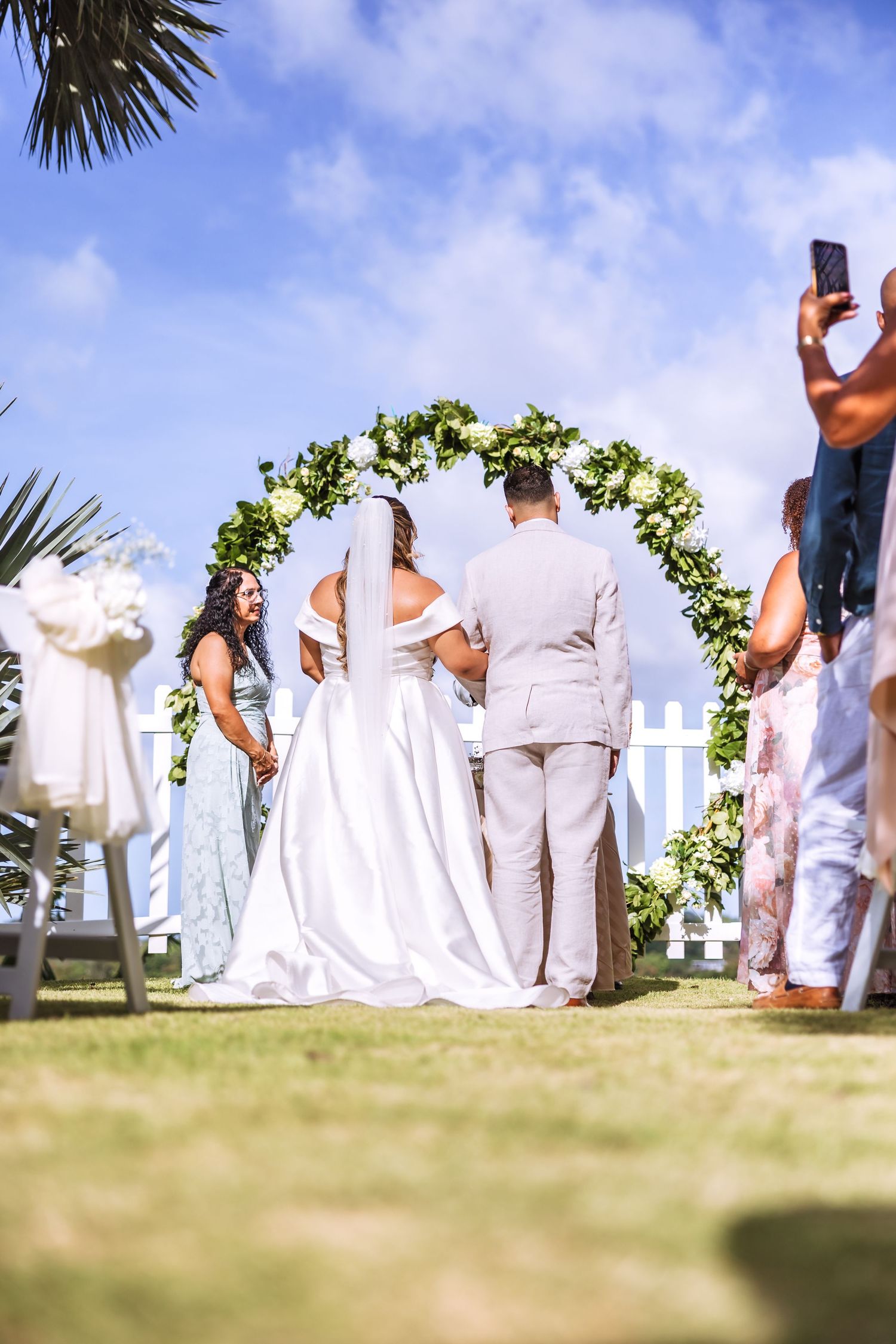 Bride and groom stand at floral arch during outdoor wedding ceremony on sunny day with guests watching.