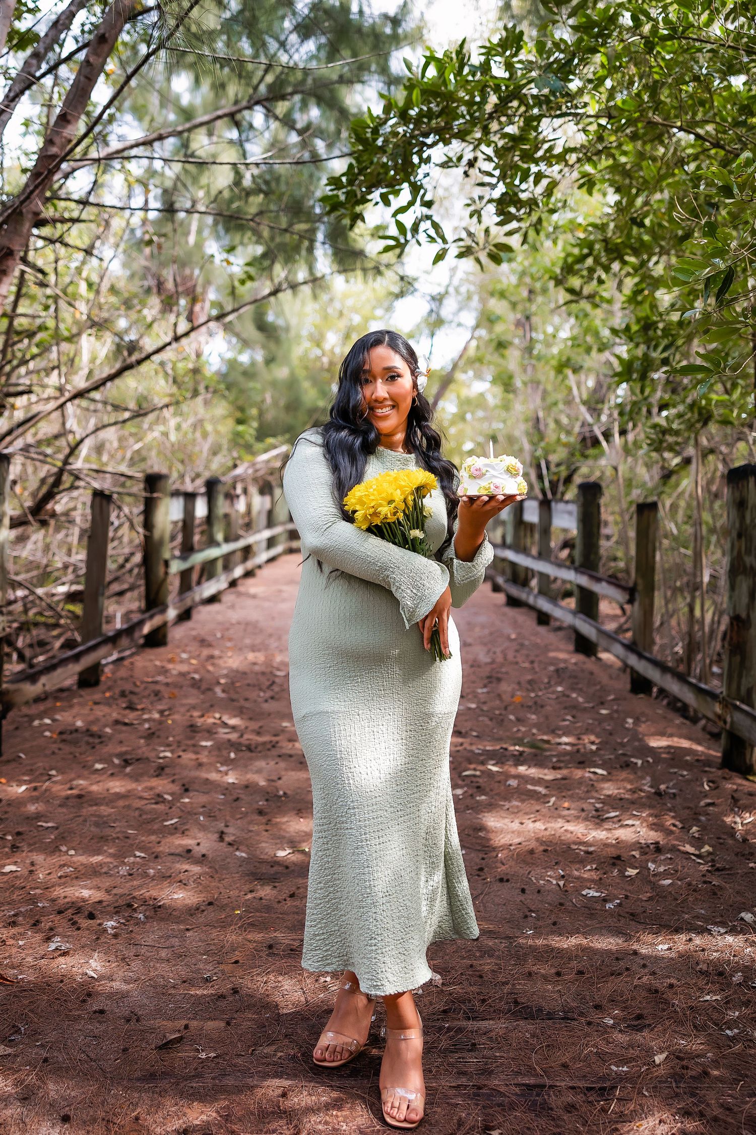 Woman in mint green dress holding yellow flowers and cake on a wooden bridge path in a forest setting.