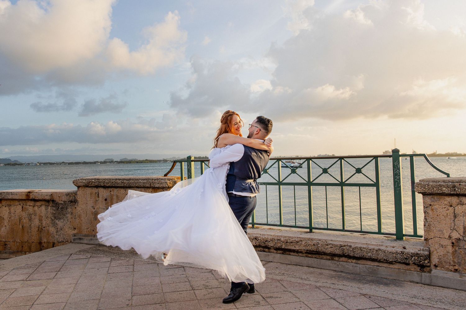 Groom lifting bride in white gown on seaside terrace at sunset, romantic wedding photo with ocean backdrop.