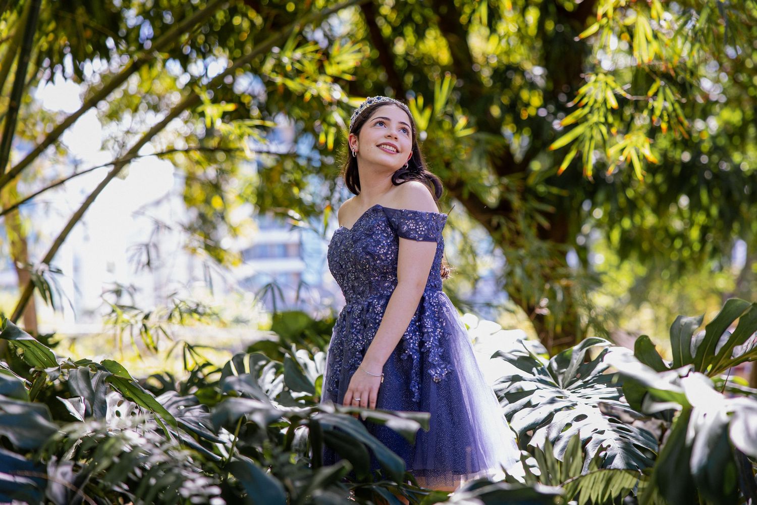 A person in a sparkly blue formal dress poses in a lush garden with tropical foliage.