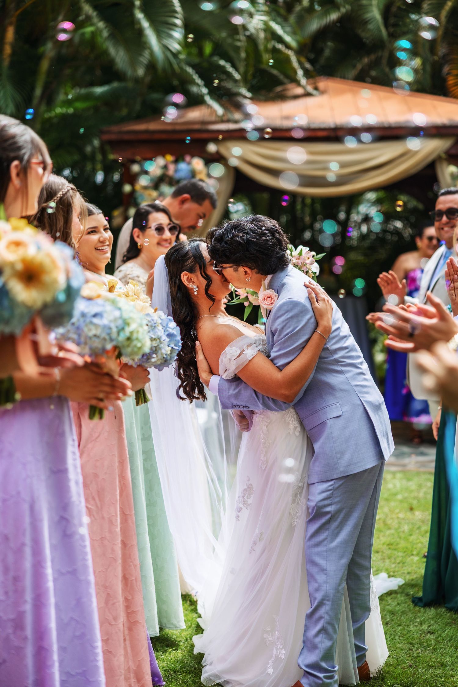 Newlyweds share their first kiss at an outdoor wedding ceremony surrounded by bridesmaids and floating bubbles.