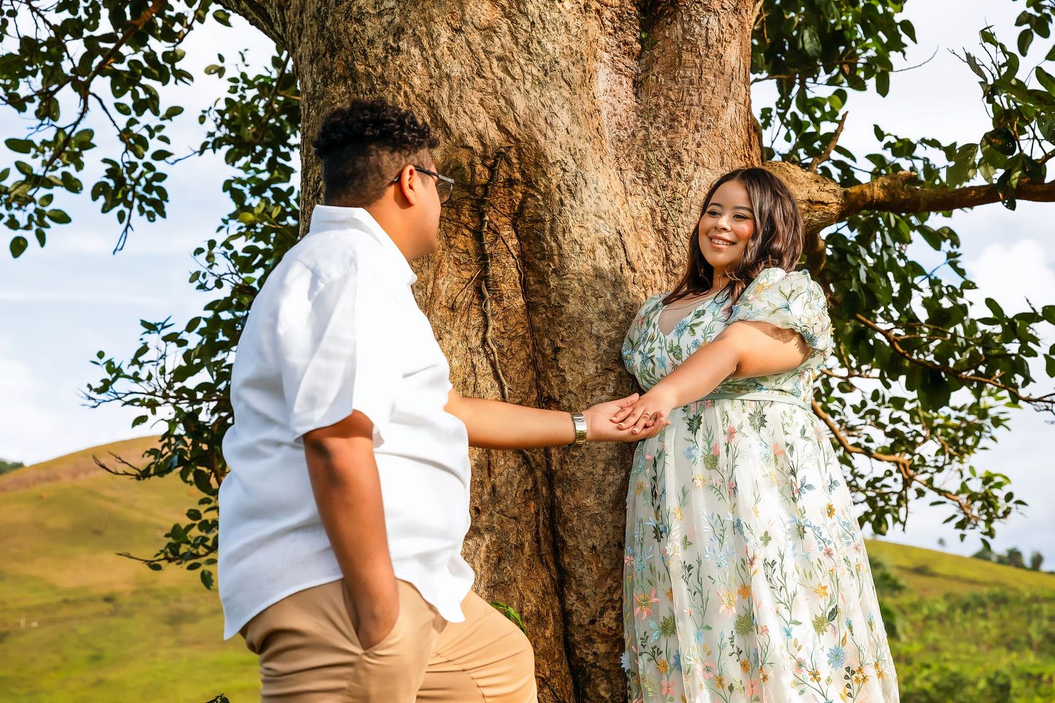 Couple holding hands and smiling near a large tree in a scenic green hillside outdoor setting.