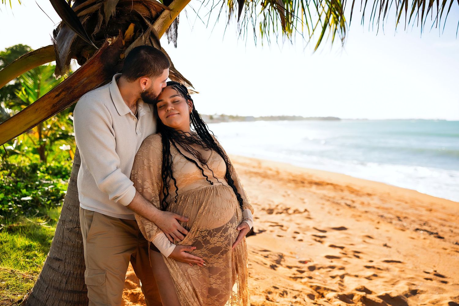 Pregnant couple embracing under a palm tree on a tropical beach at golden hour.