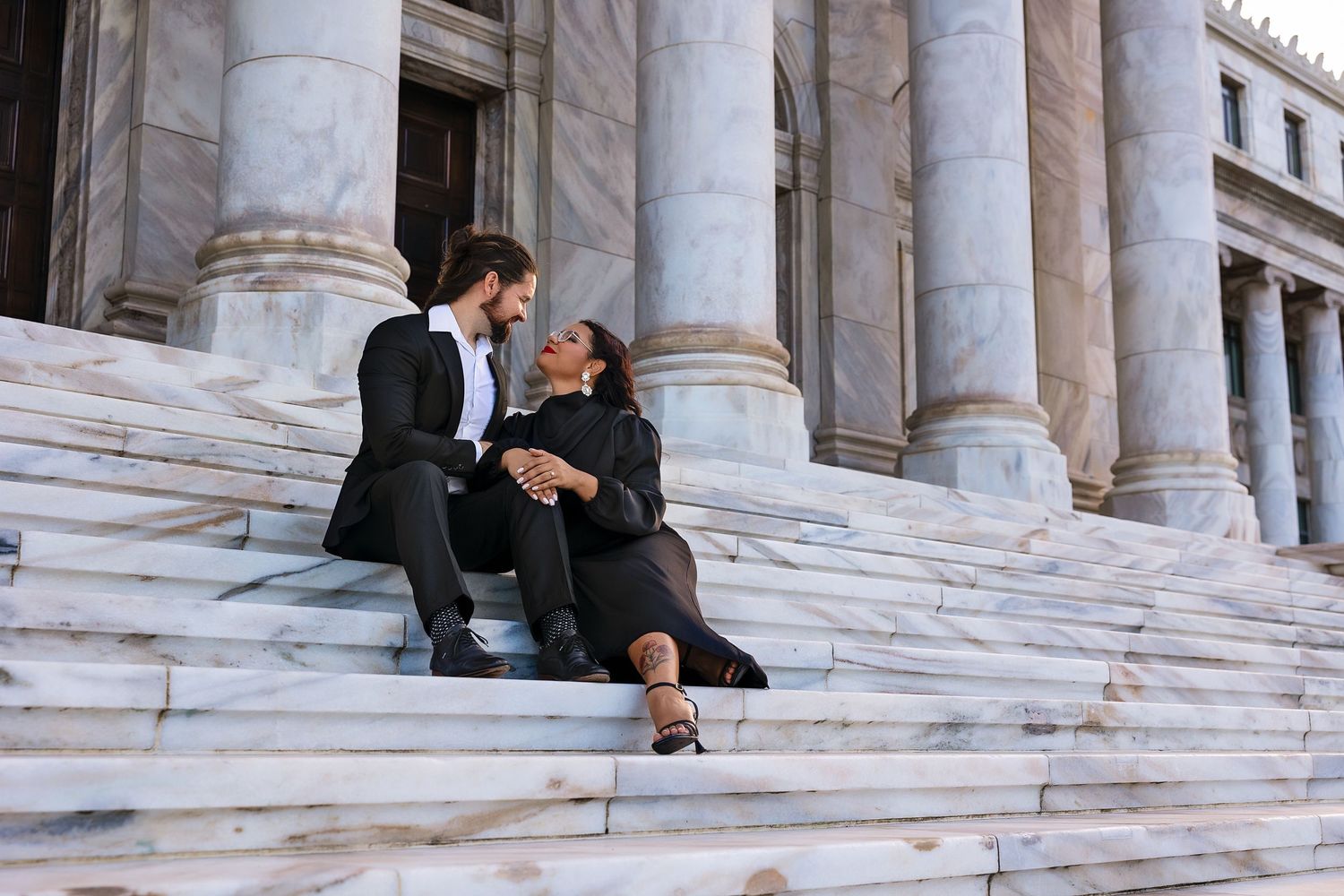 Couple in formal attire sitting intimately on marble steps of a grand building with tall columns.
