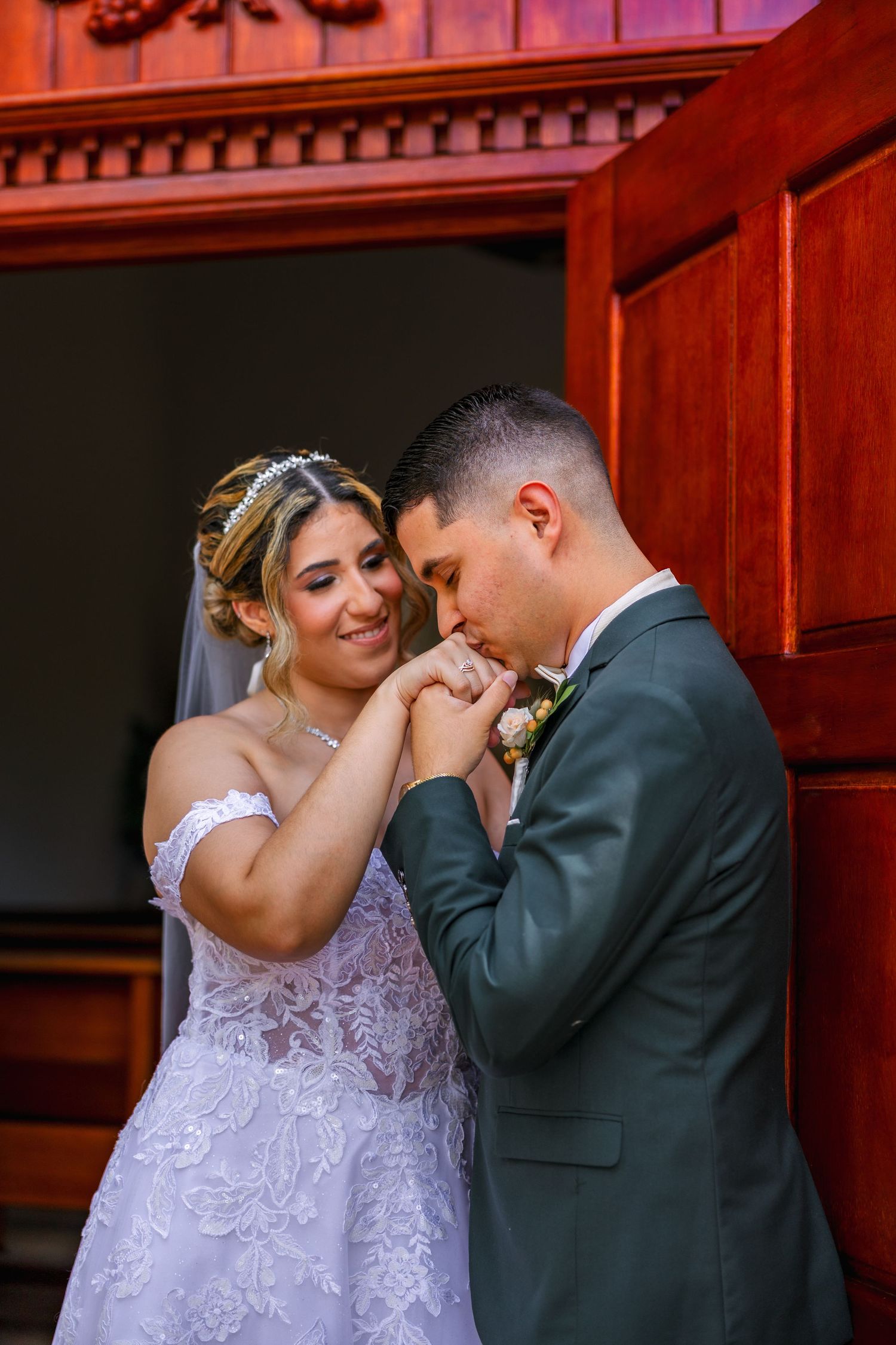 Groom in gray suit kissing bride's hand while she smiles, standing by ornate wooden doors in wedding attire.