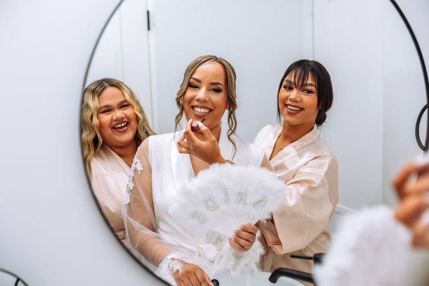 Three women in white robes smile and laugh together while getting ready in front of a bathroom mirror.