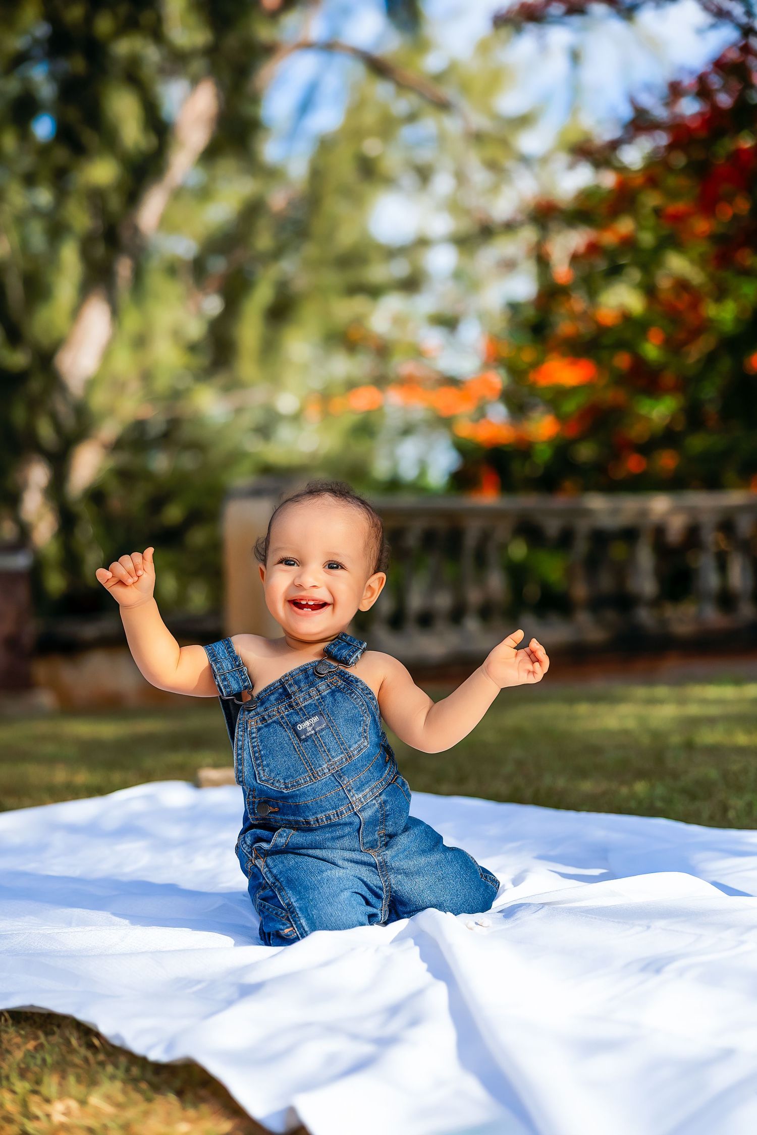 Happy baby in denim overalls sitting on white blanket outdoors, smiling with arms raised in sunny garden setting.