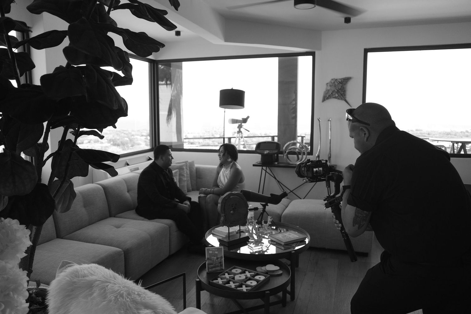 Black and white photo of a film crew interviewing people on a modern white sofa in a bright living room.