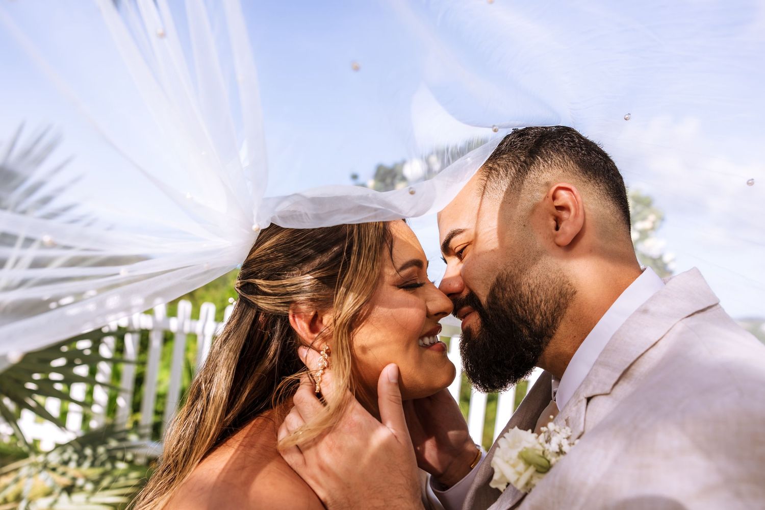 A bride and groom share a romantic almost-kiss under a flowing white veil outdoors on their wedding day.