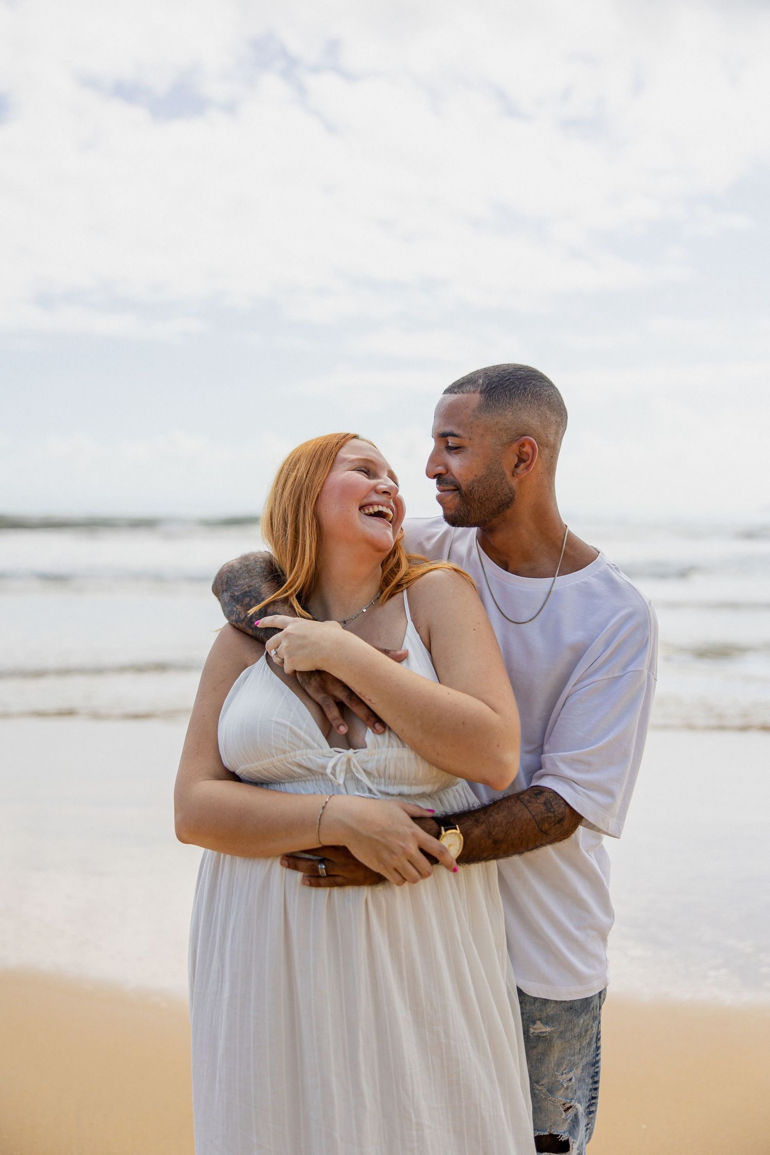 Happy couple embracing on a sandy beach, woman in white dress holding a puppy, smiling at each other with ocean waves behind.