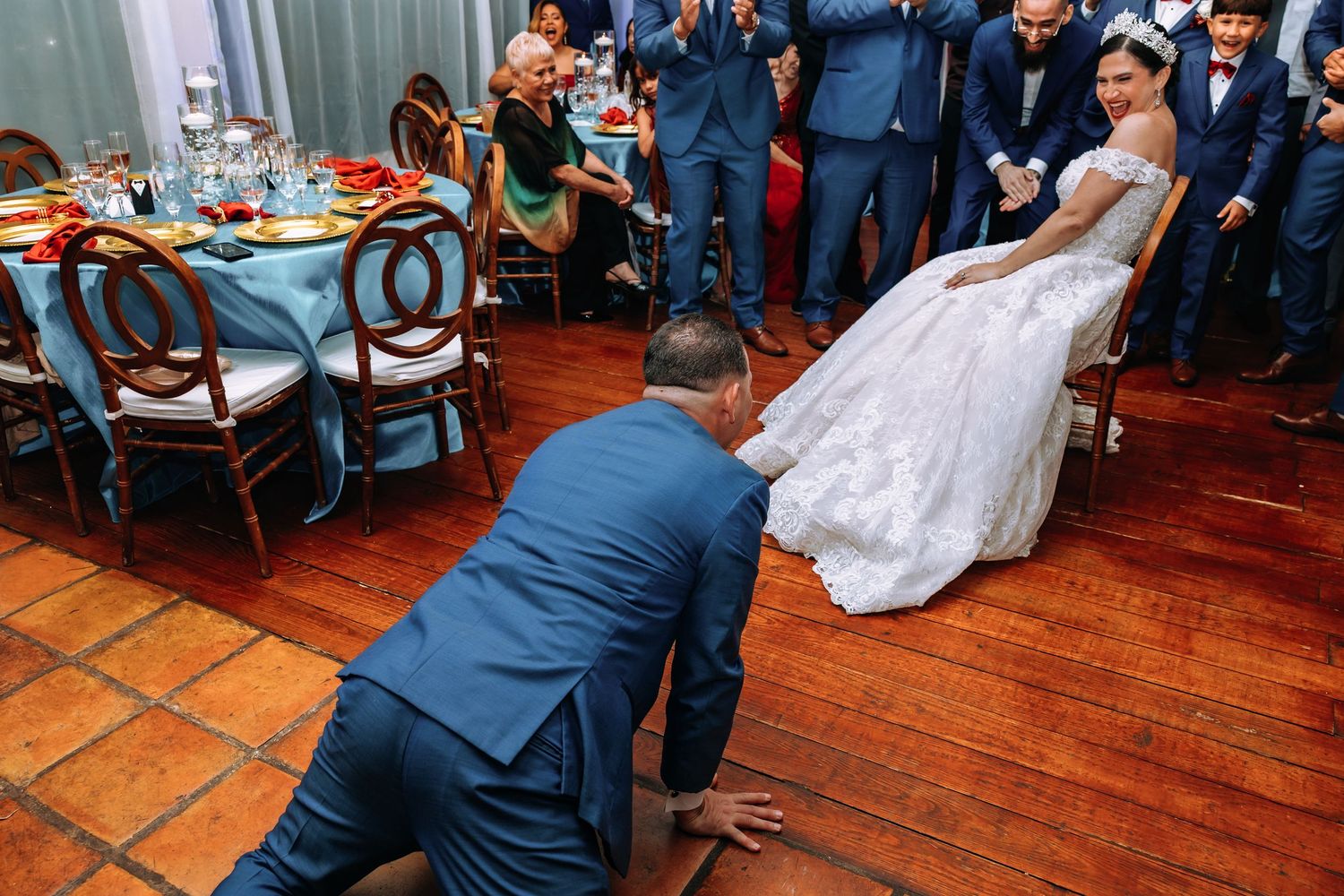 Groom kneeling before bride in white dress during wedding reception on wooden dance floor.