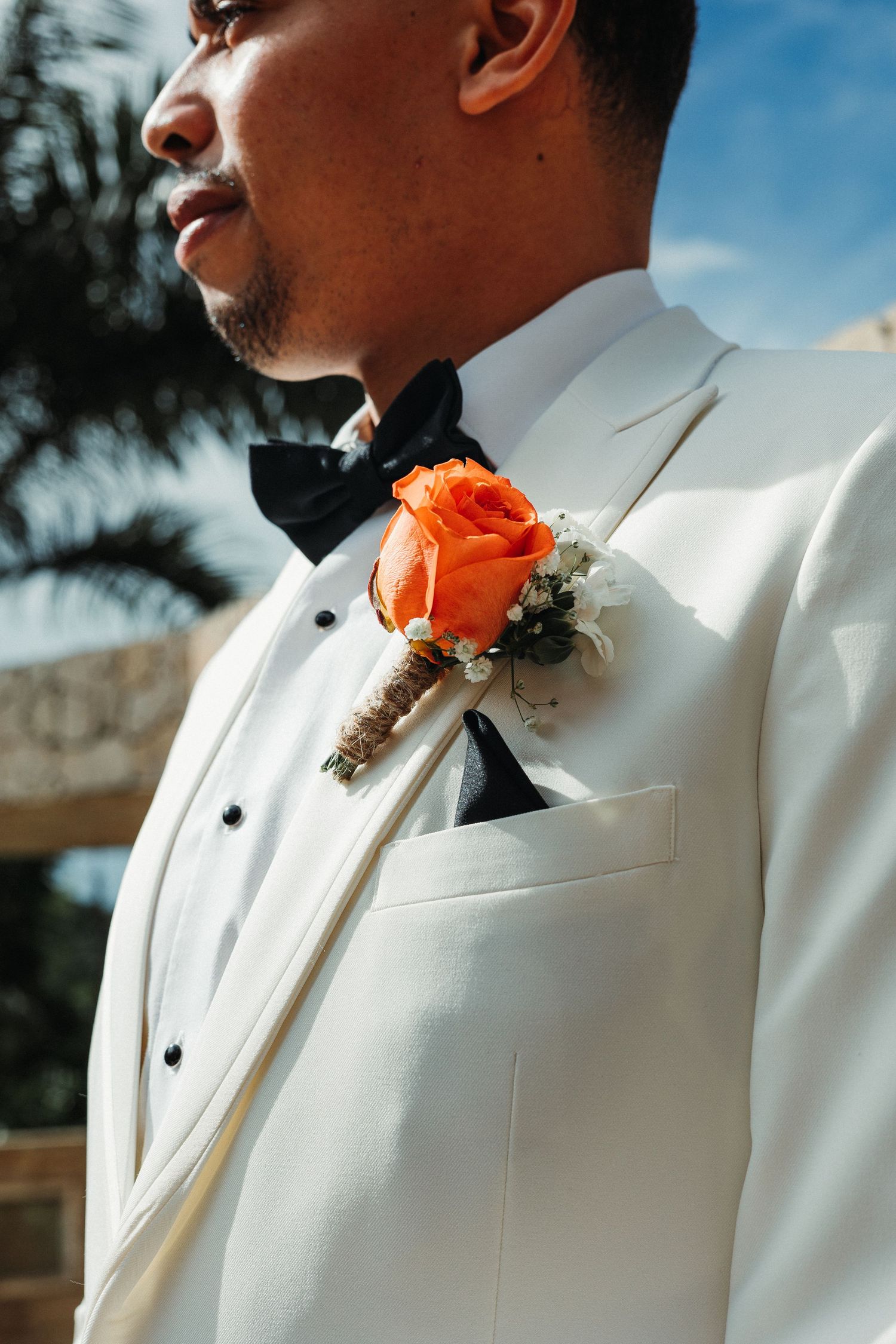 Groom in white tuxedo with black bow tie and orange rose boutonniere at outdoor wedding ceremony.