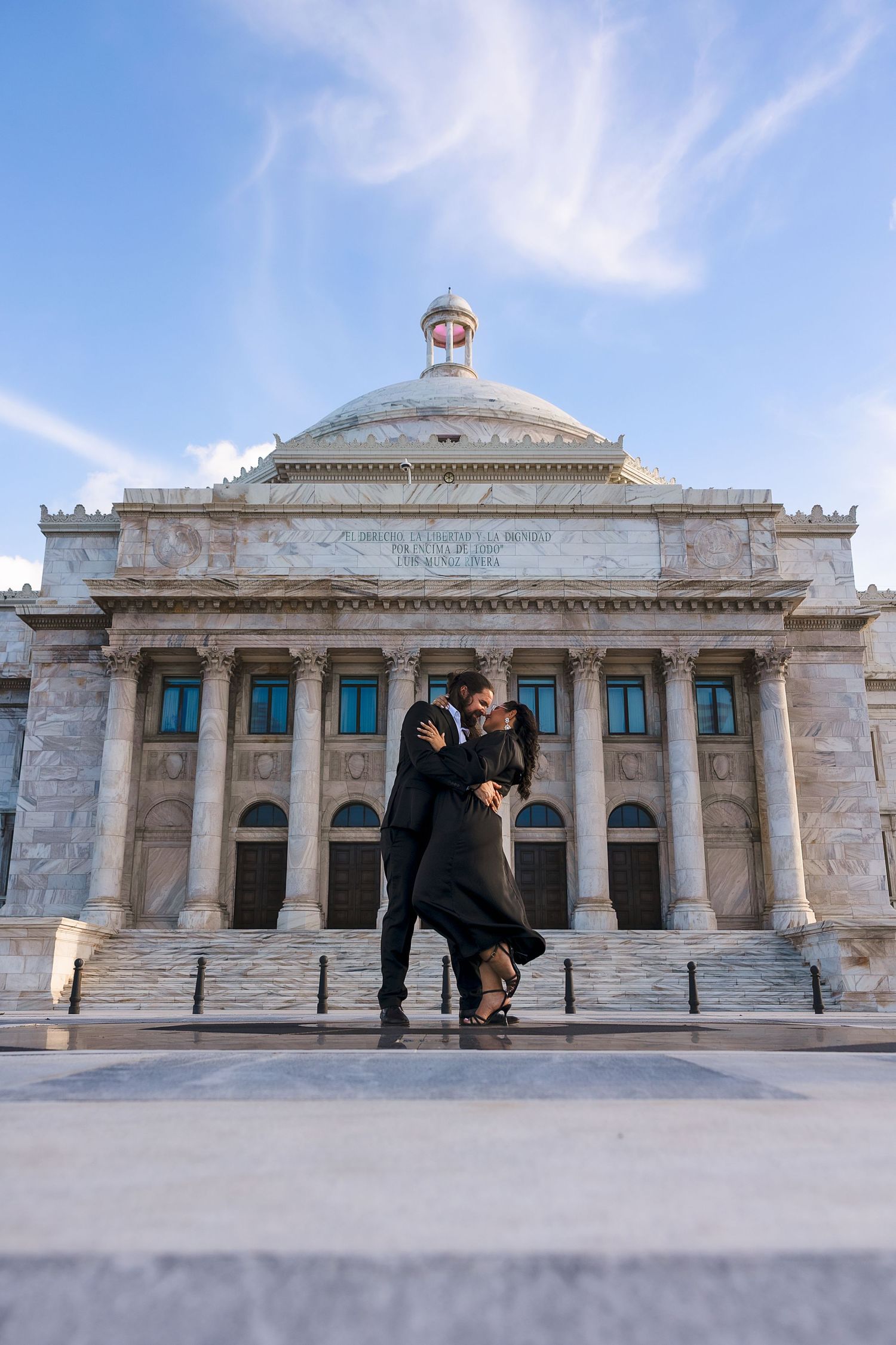 A romantic couple dancing in front of a neoclassical building with a domed roof and columns on a sunny day.