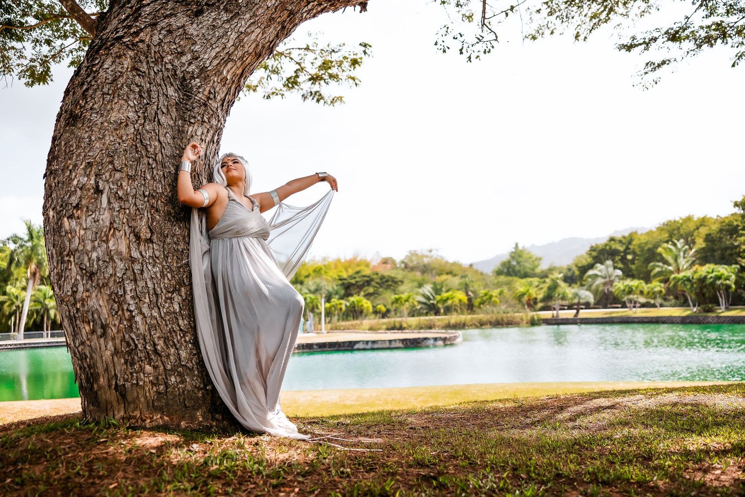Woman in flowing gray dress poses gracefully against large tree trunk beside serene green lake in tropical park.
