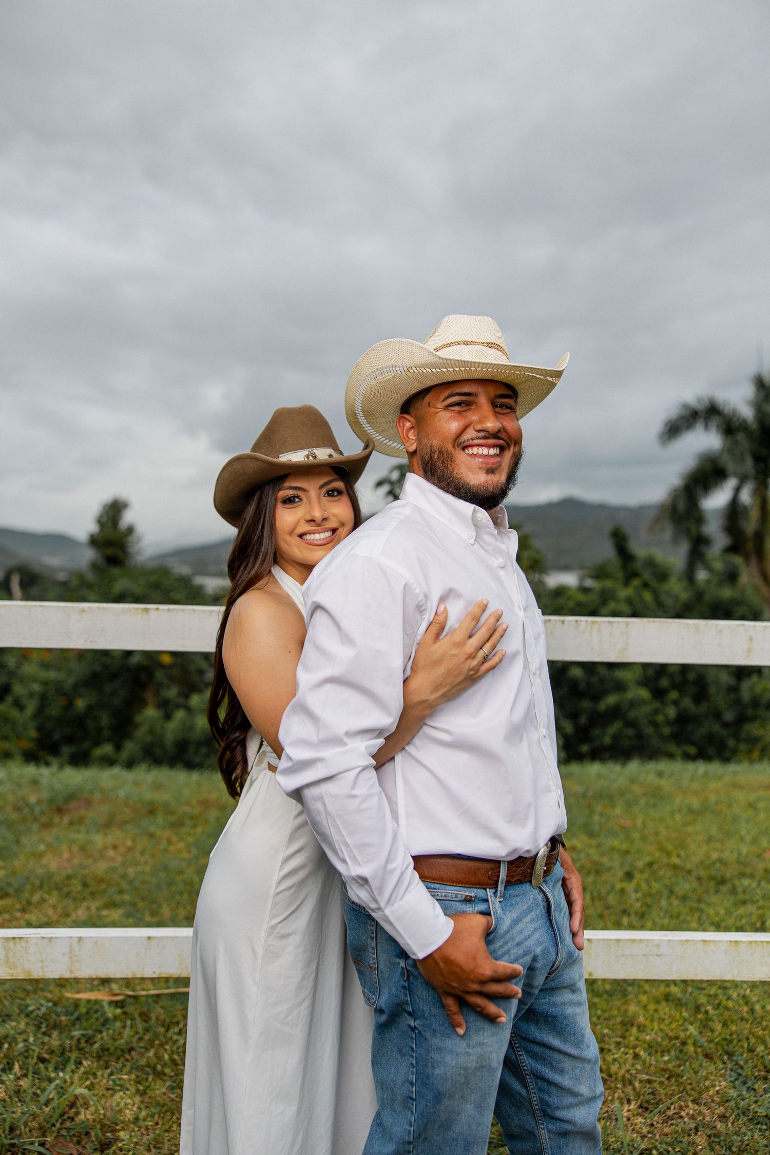 Smiling couple in cowboy hats posing by a white fence in a green rural landscape.