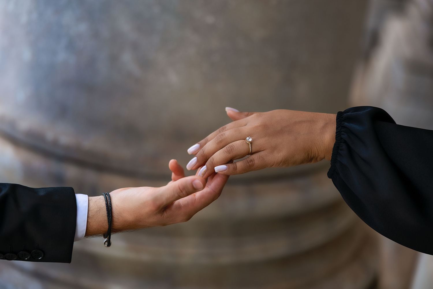 Two hands gently touching, one wearing a diamond engagement ring, against a blurred metallic background.