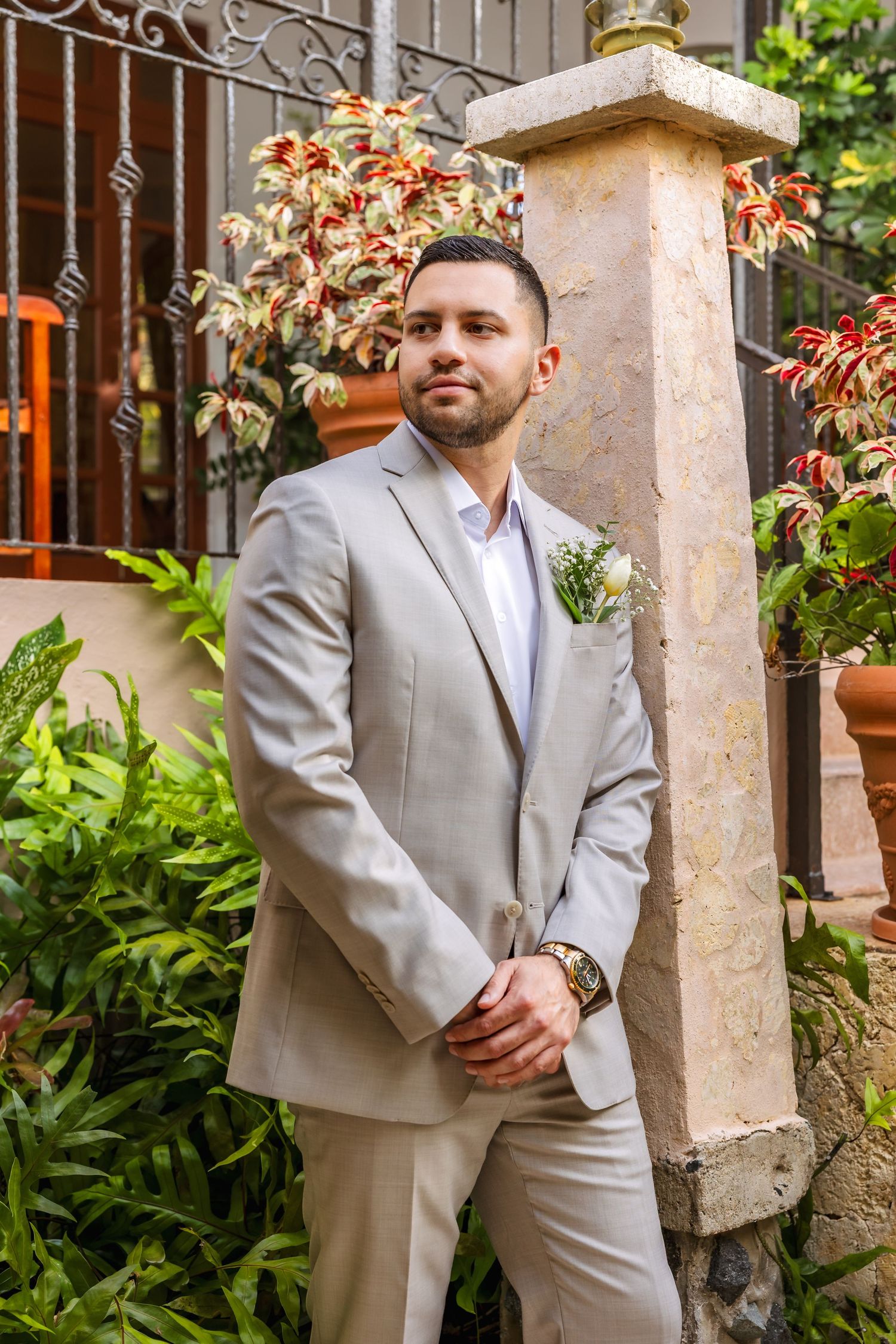 Groom in a tan suit with boutonniere poses by a stone pillar surrounded by lush tropical greenery.