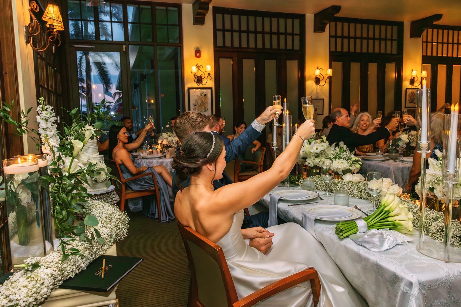 Bride raises a champagne toast at an elegant indoor wedding reception with white floral arrangements and candlelit tables.