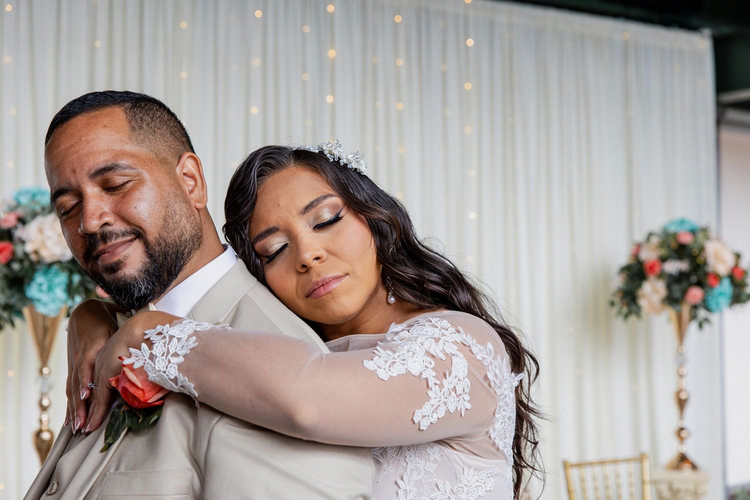 Bride in lace gown rests head on groom's shoulder during emotional wedding dance at decorated reception venue.