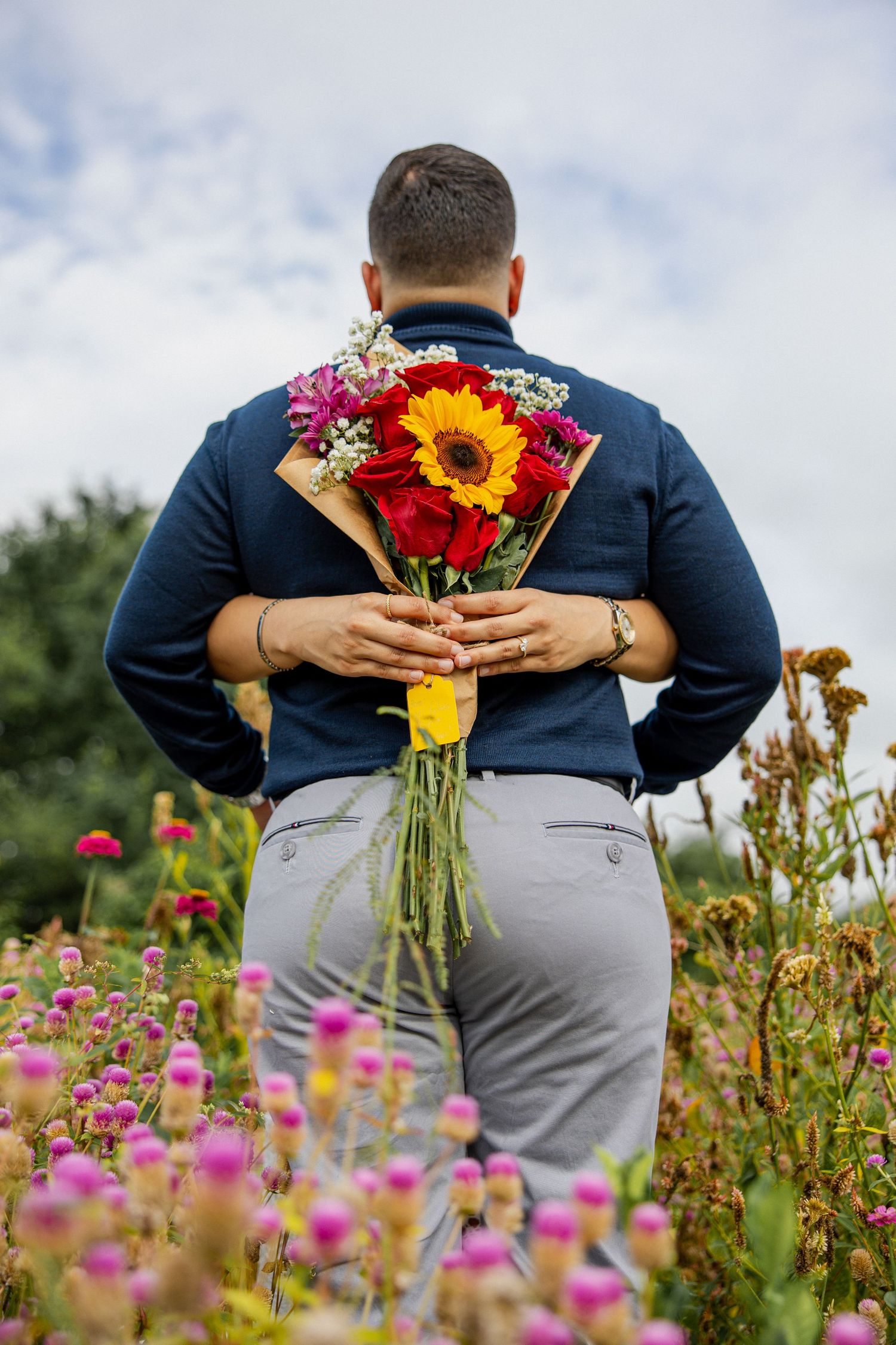 Man holding colorful bouquet with sunflower behind his back while standing in a vibrant flower field.