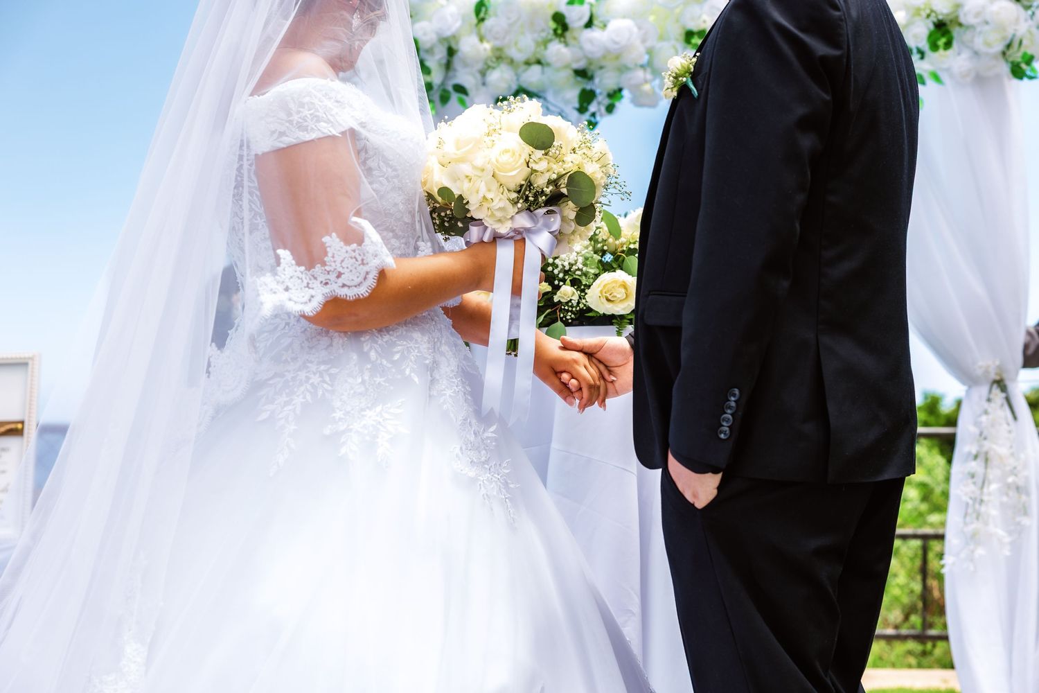 Bride and groom holding hands at outdoor wedding ceremony, bride wearing lace ball gown holding white floral bouquet.