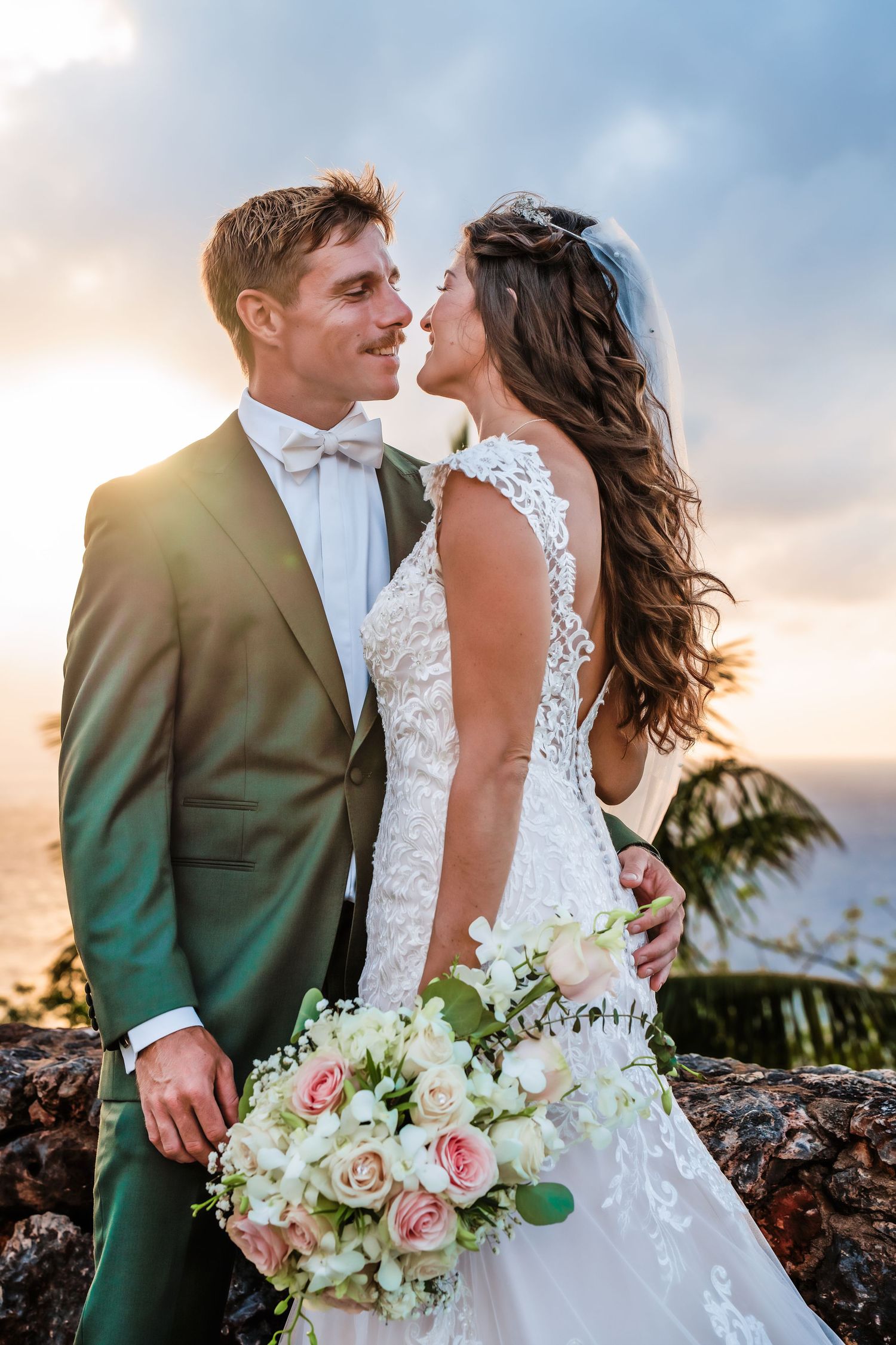 Bride and groom share a romantic moment at sunset, she holds a bouquet of pink and white roses, both dressed in elegant wedding attire.