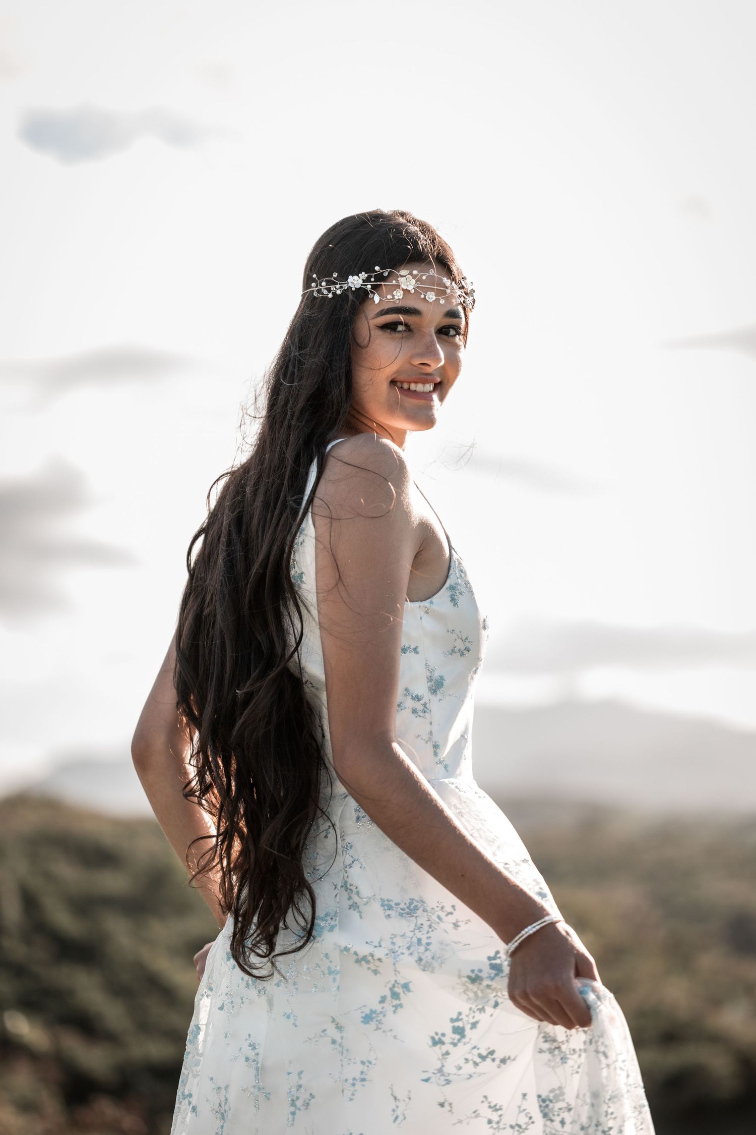 Smiling bride in white floral dress with long dark hair and floral crown poses outdoors against a bright sky.