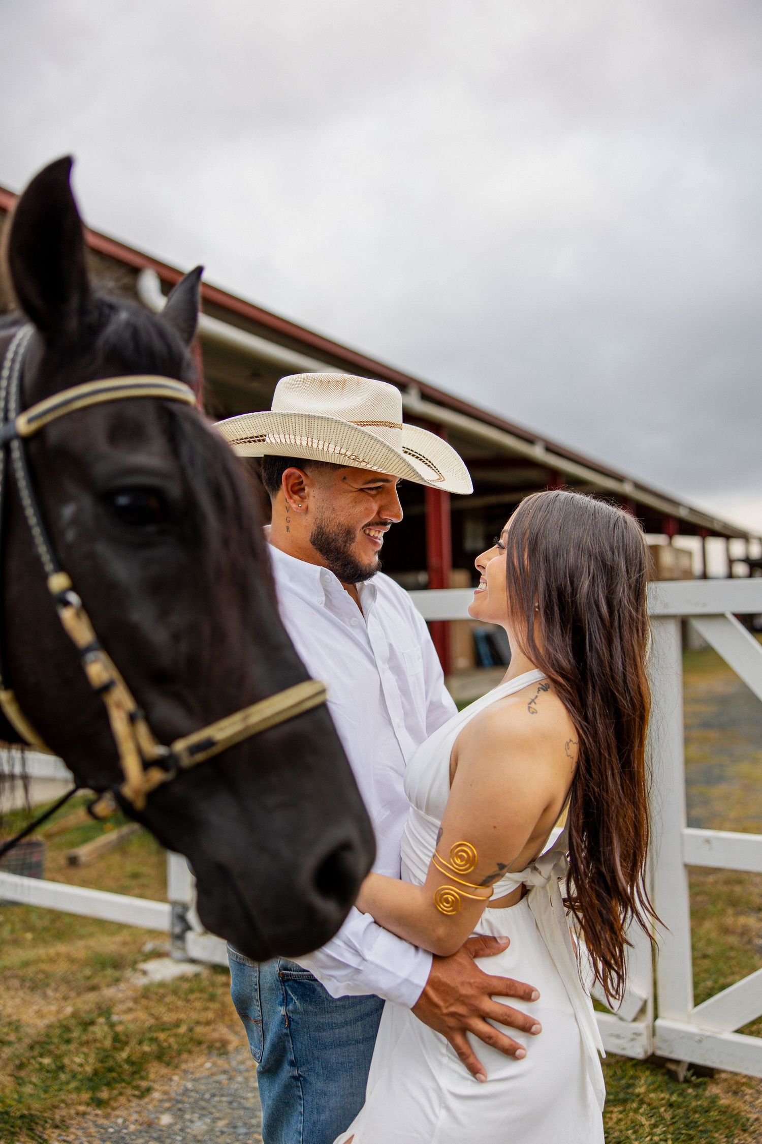 Couple embracing near a black horse at a ranch, man in cowboy hat and white shirt, woman in white dress.