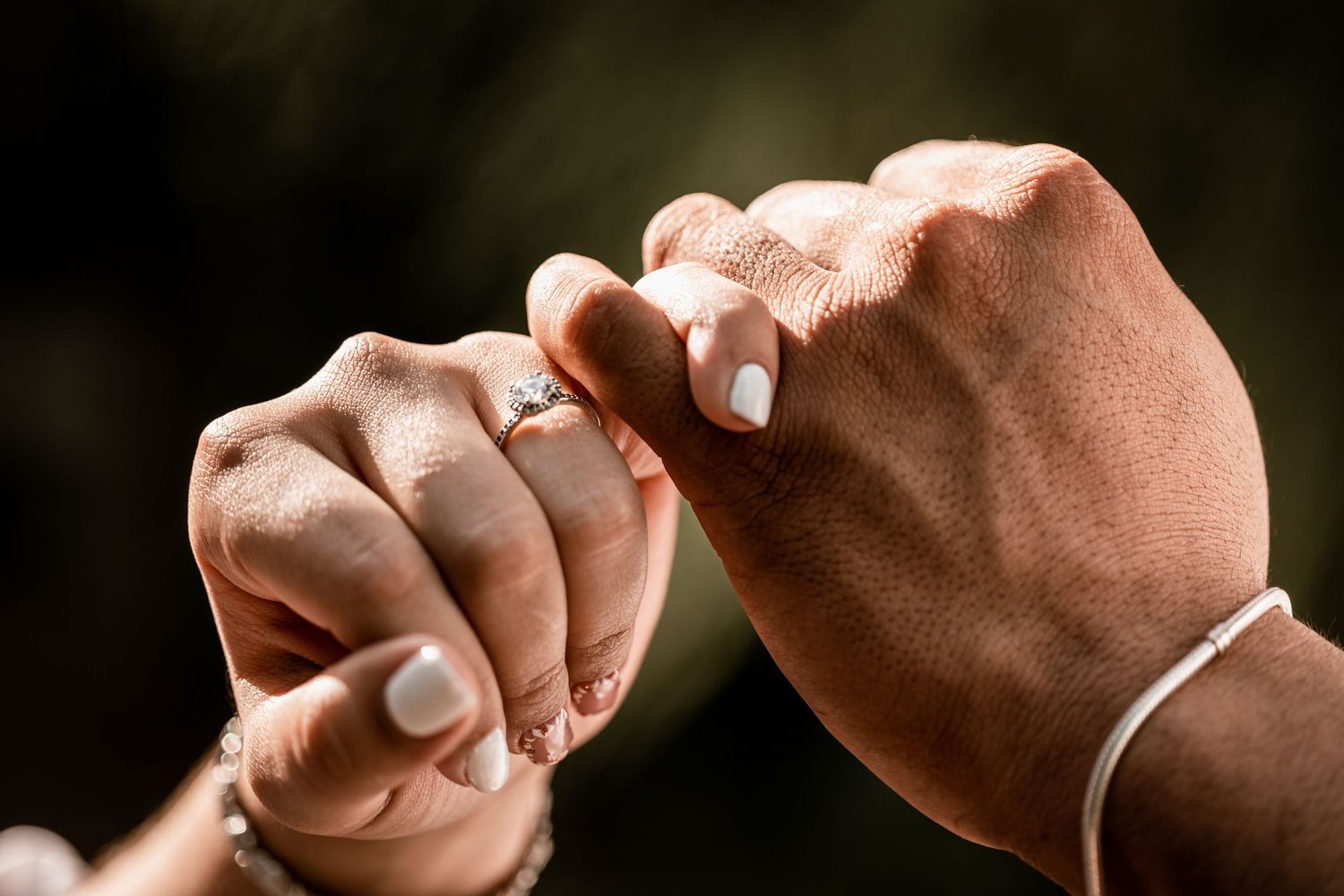 Two hands with white manicured nails and a diamond engagement ring gently linking pinky fingers together.