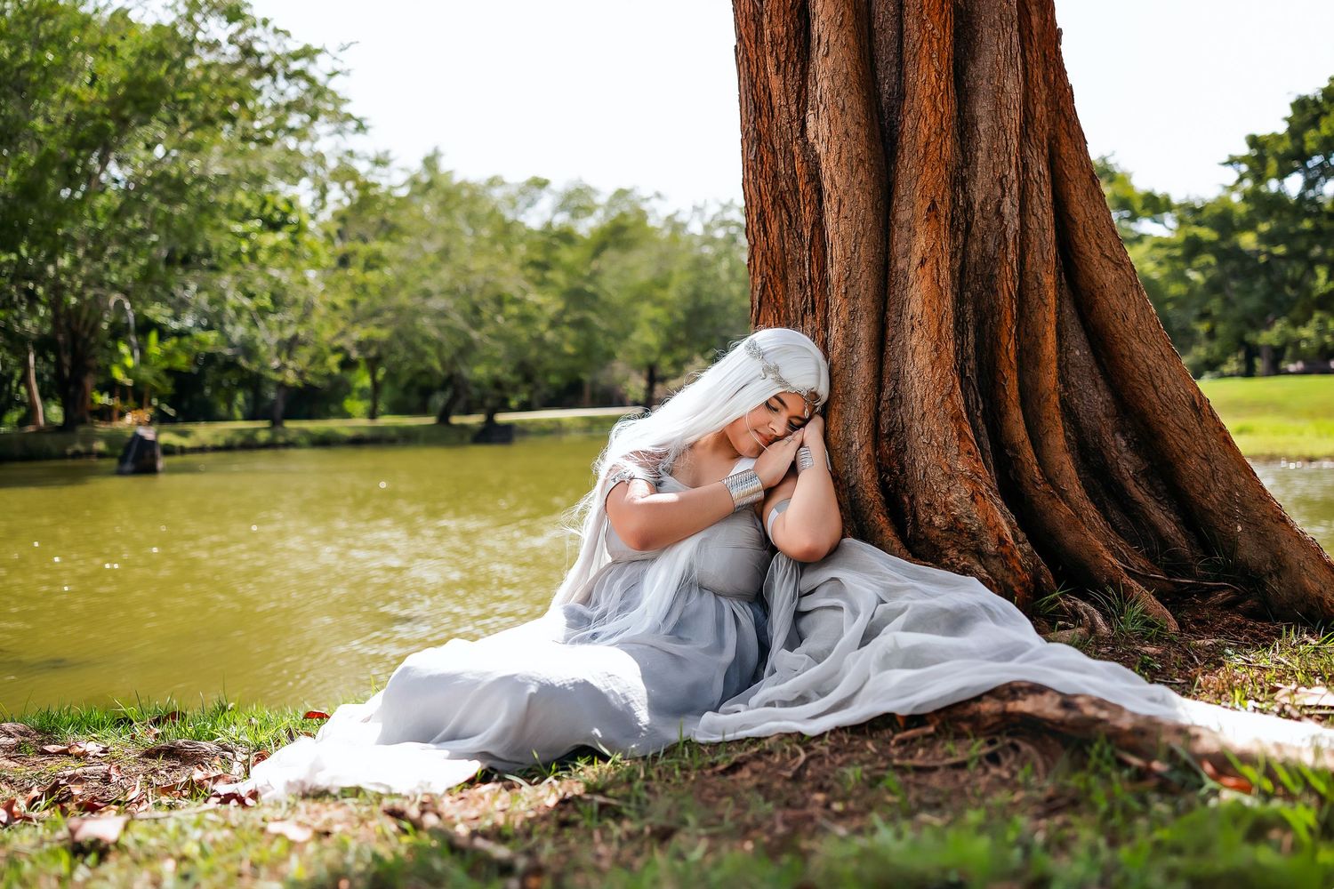 Woman in white dress resting peacefully against large tree trunk by serene lake in lush green park.