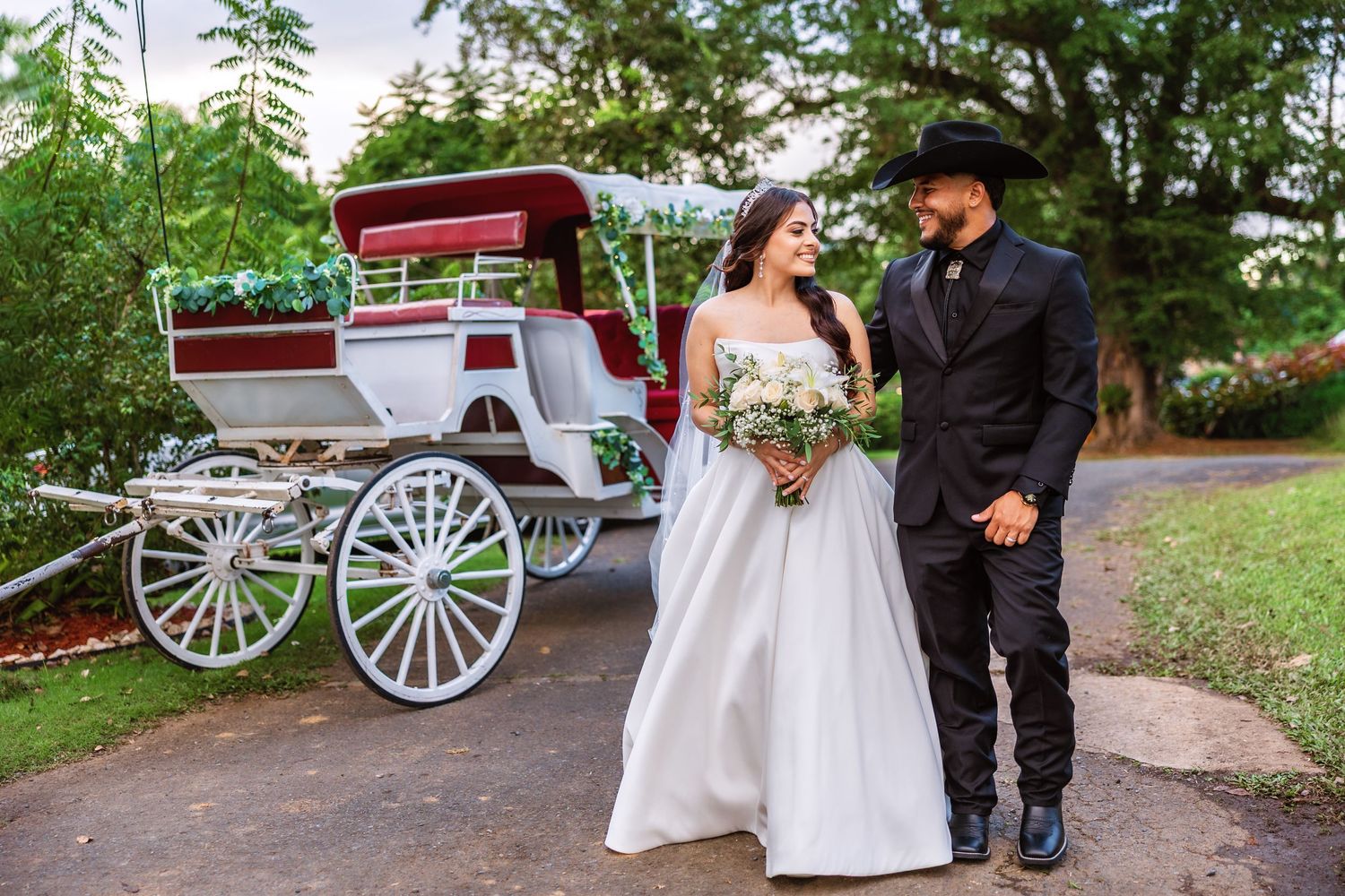 Bride in white gown and groom in black suit stand beside a white horse-drawn carriage in a lush garden setting.