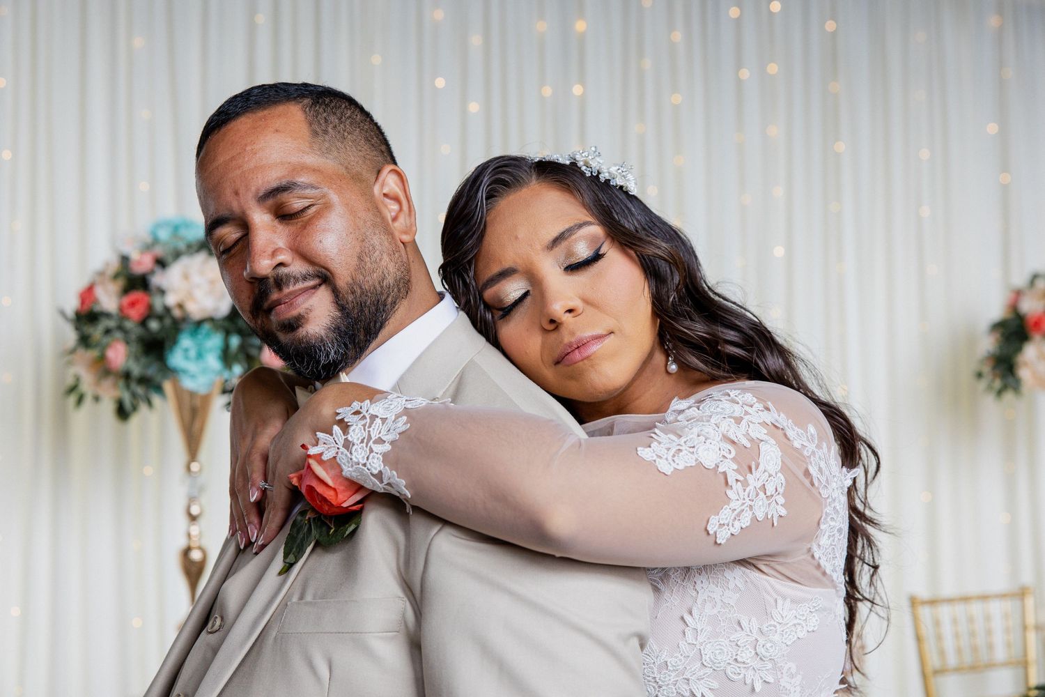 Newlyweds share an intimate moment at their wedding reception with floral decorations and white backdrop.