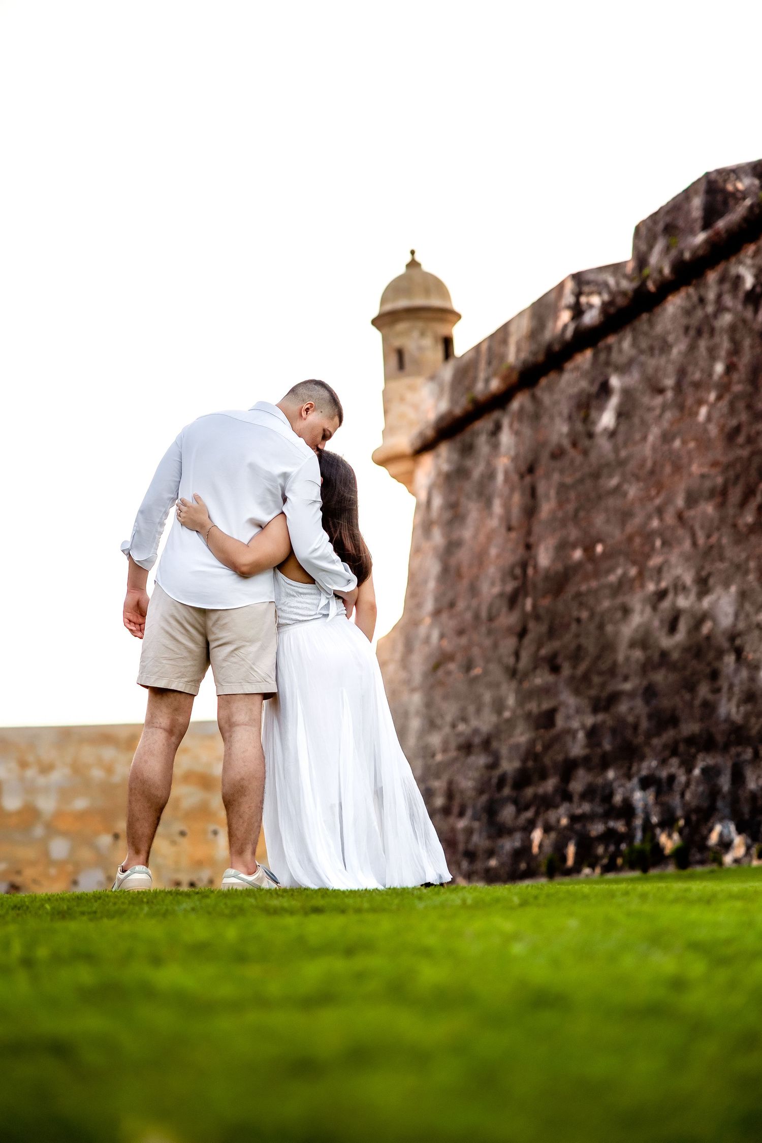 A couple in white clothing poses together on a grassy lawn near an old stone fortress wall.