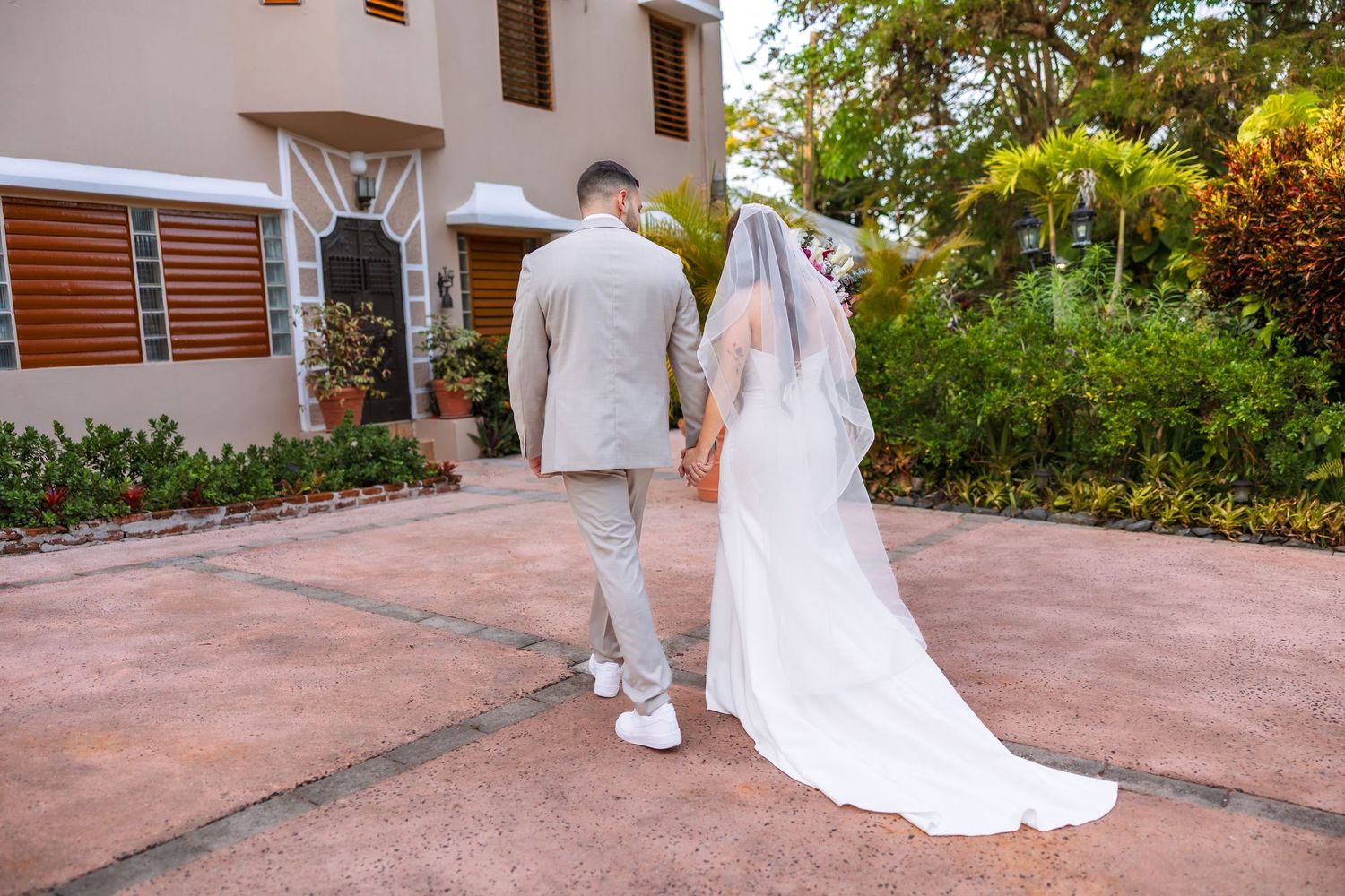 Bride and groom walking hand in hand away from camera, wearing white wedding dress with veil and beige suit.