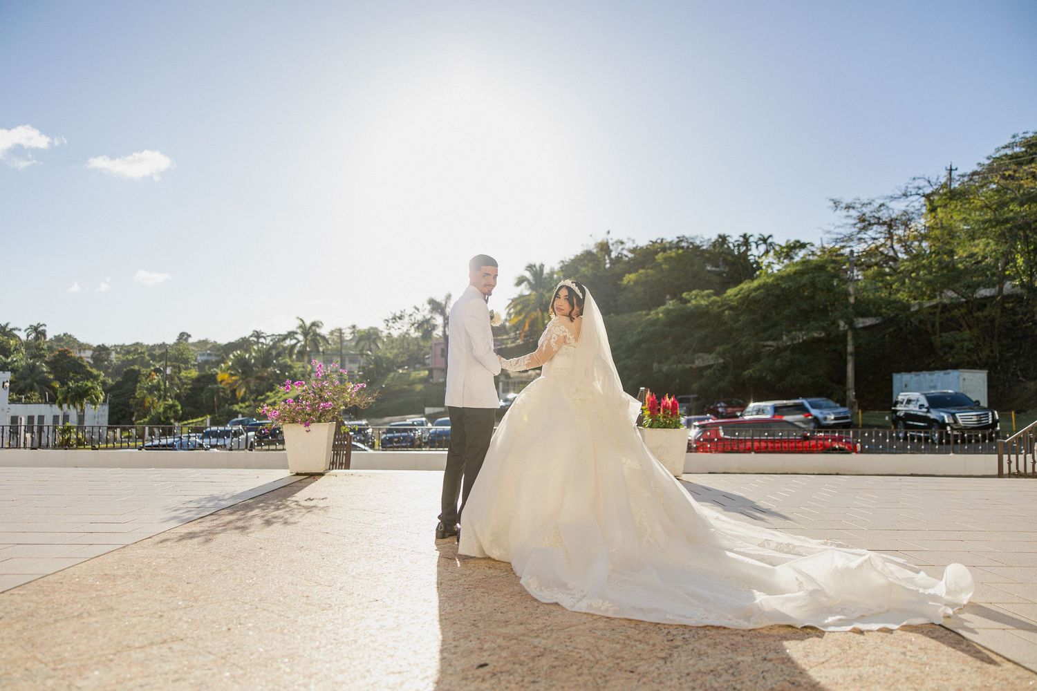 A couple in wedding attire stands together on a sunlit waterfront overlooking a harbor with palm trees in the background.