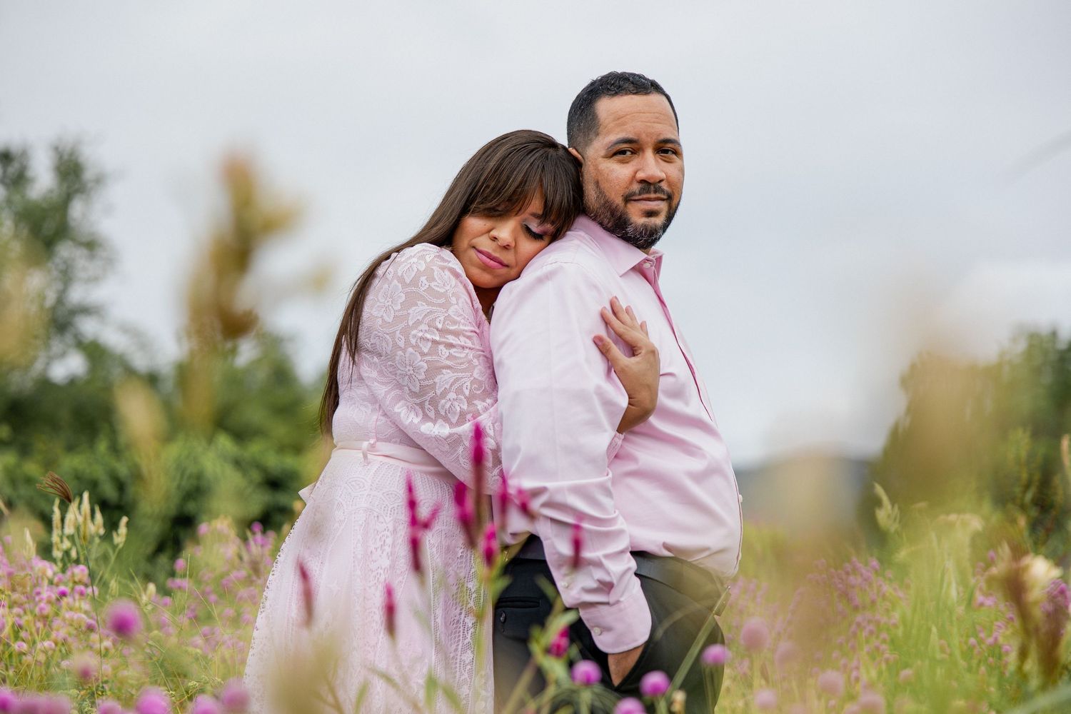 A couple embraces in a field of pink wildflowers on a cloudy day.