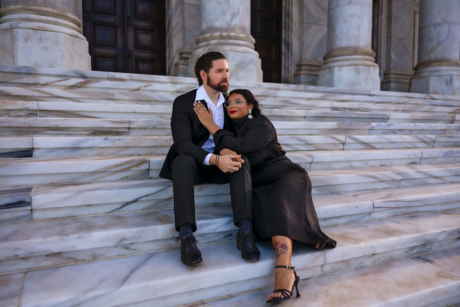 Two people sitting and embracing on marble steps of a grand building in formal attire.
