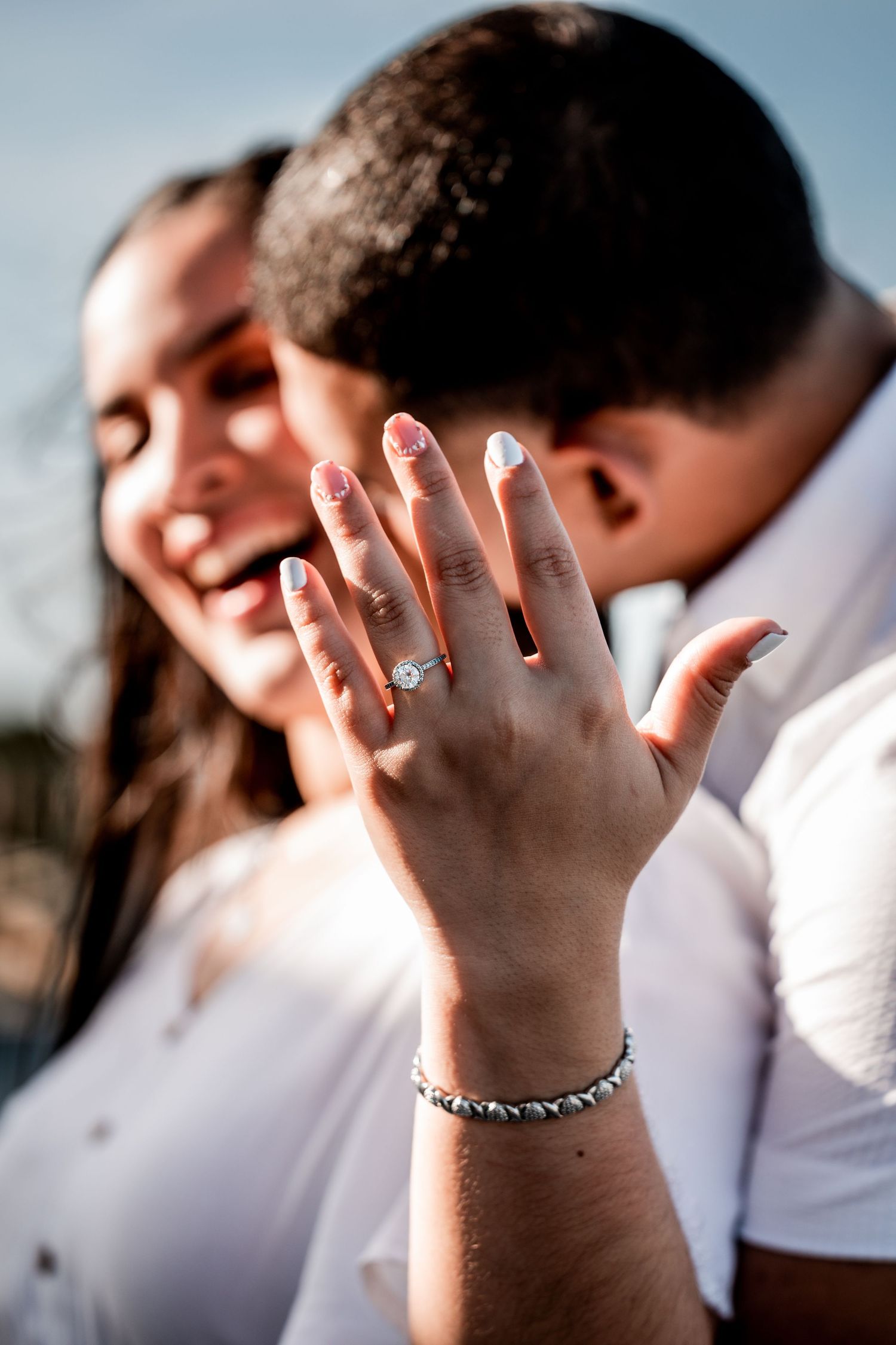 Happy couple in white shirts showing off diamond engagement ring, woman smiling with manicured nails and bracelet.