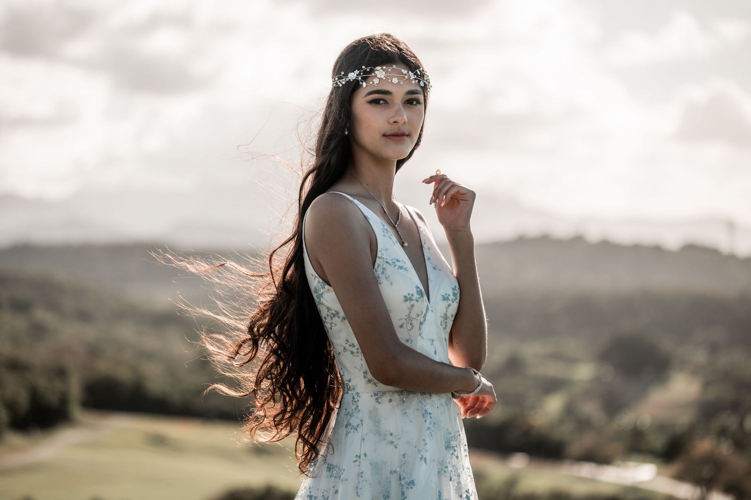 Young woman in floral dress and headpiece poses outdoors with wind-blown hair against scenic landscape backdrop.