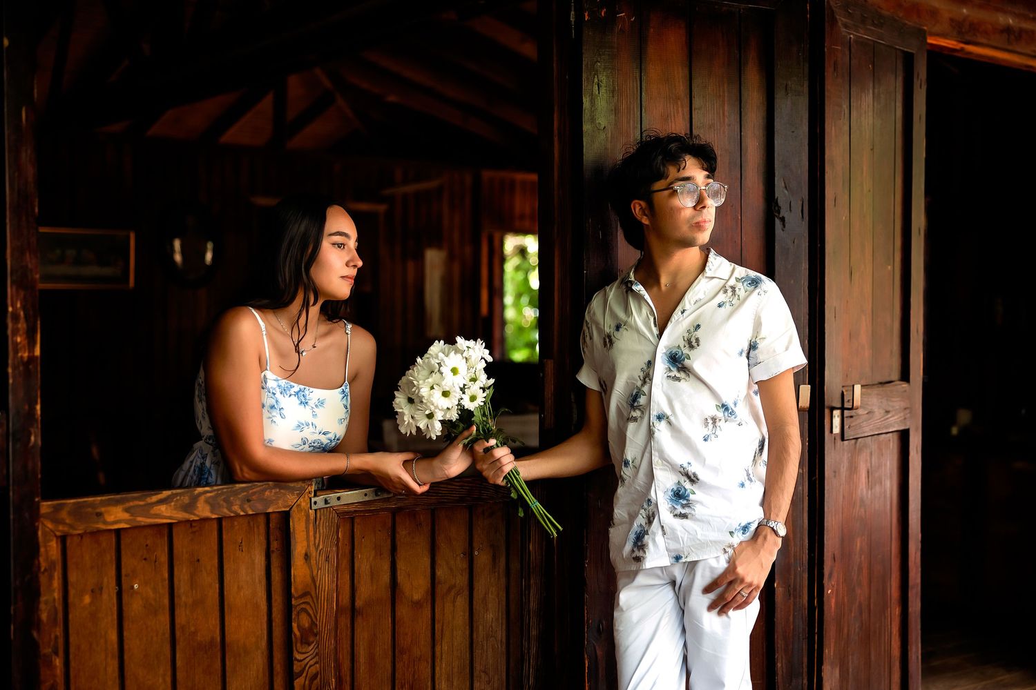 Young couple standing in rustic wooden barn doorway, woman holding white flower bouquet.