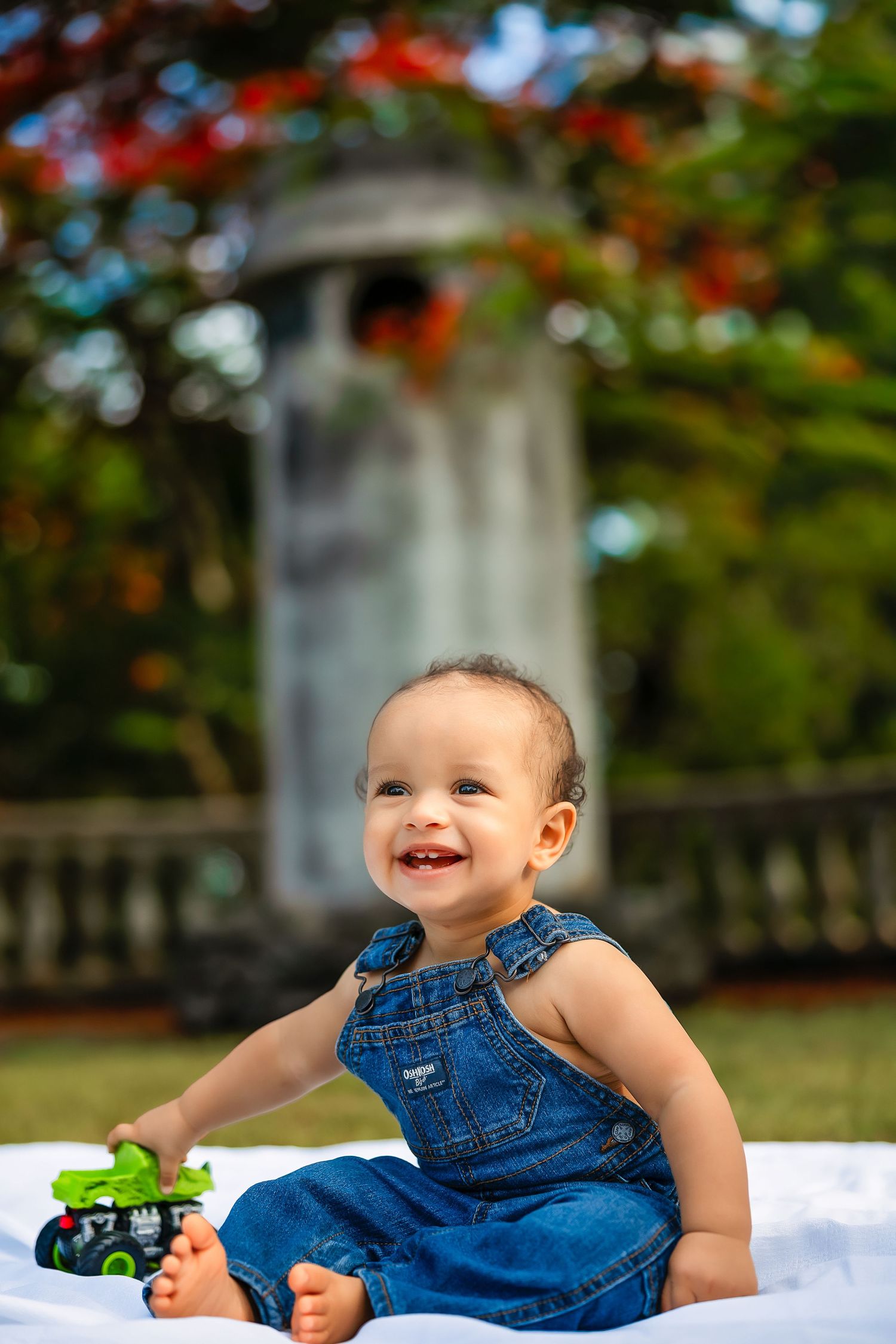 Happy baby in denim overalls sitting on white blanket outdoors, smiling with toy, garden gazebo and red flowers in background.