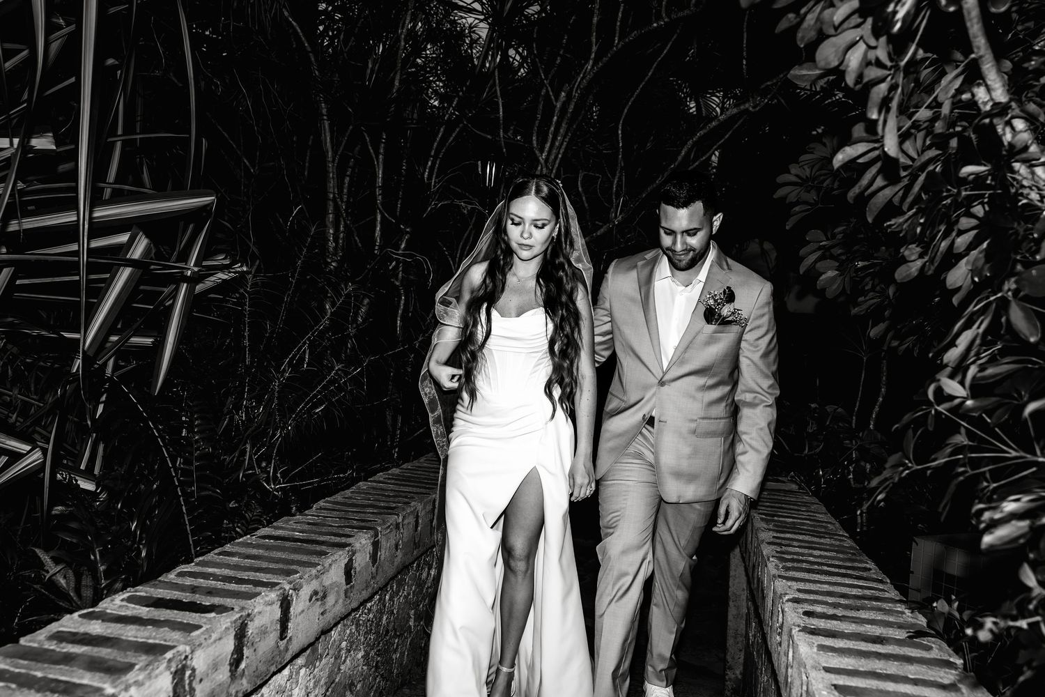 Black and white photo of a bride and groom walking hand in hand on a bridge surrounded by lush tropical greenery.