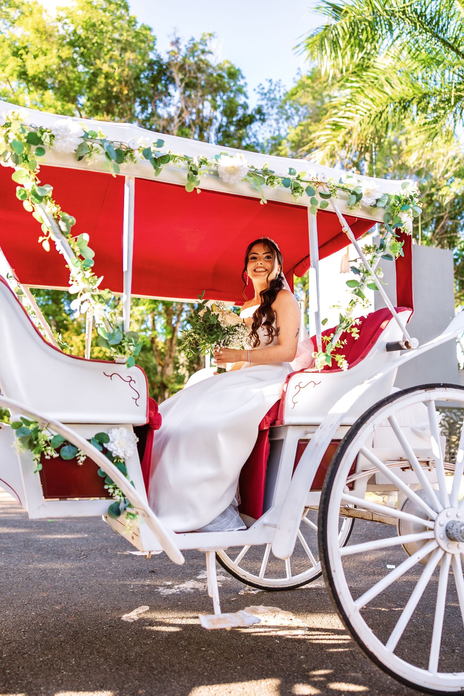 Smiling bride in white gown sitting in decorated white and red horse carriage with greenery garlands outdoors.